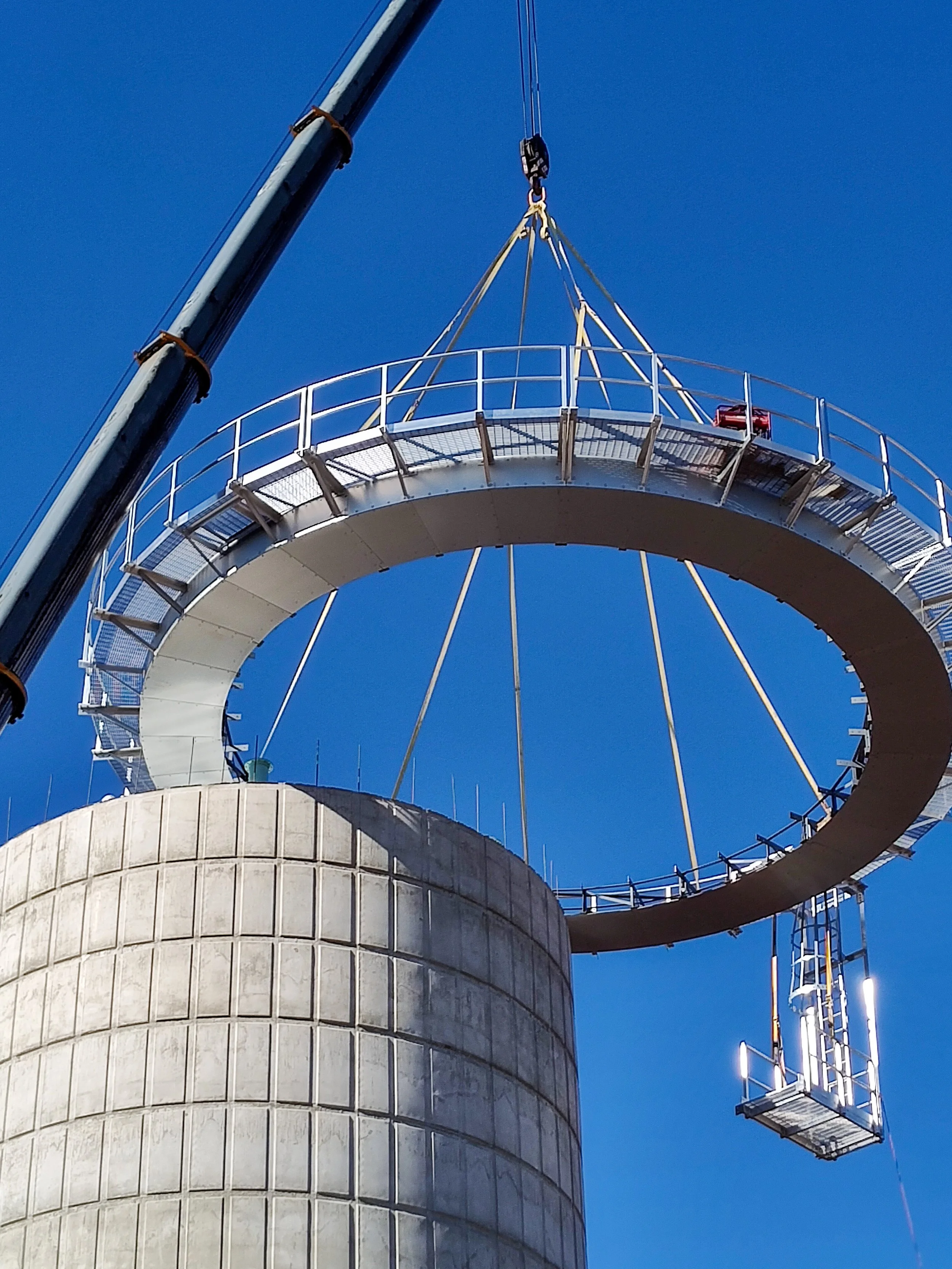 A crane lifting a large circular metallic platform or ring over a concrete silo against a clear blue sky.