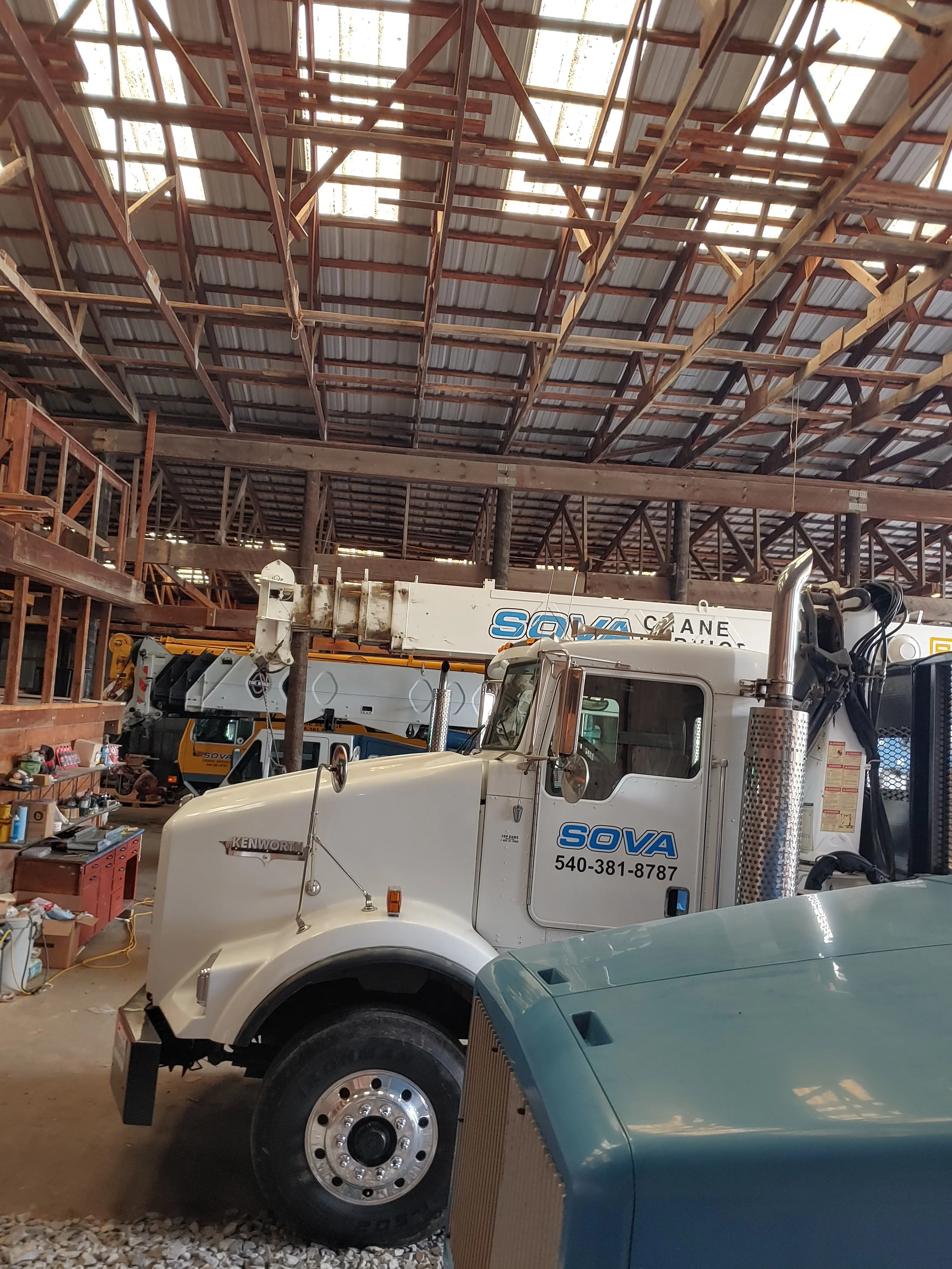 The image shows a large white tow truck with the company name 'SOVA' and phone number on the side, parked inside a spacious warehouse with a high, partially open ceiling and exposed wooden beams. There is an assemblage of tools and shelves to the lef