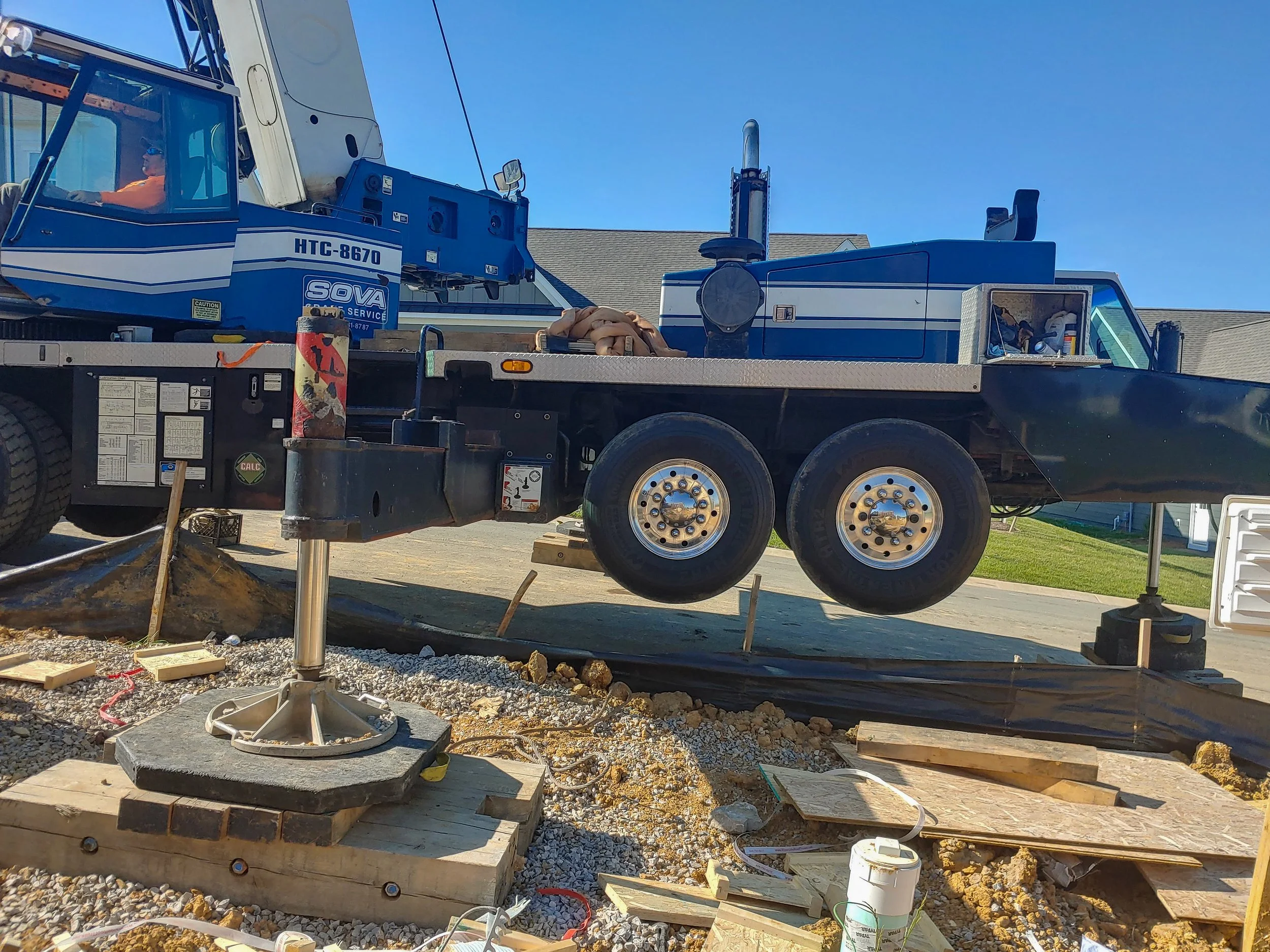 A construction site with a black flatbed truck. The truck is suspended in the air by a hydraulic lift, with construction materials and debris on the ground below, including wood planks, concrete blocks, and tools.