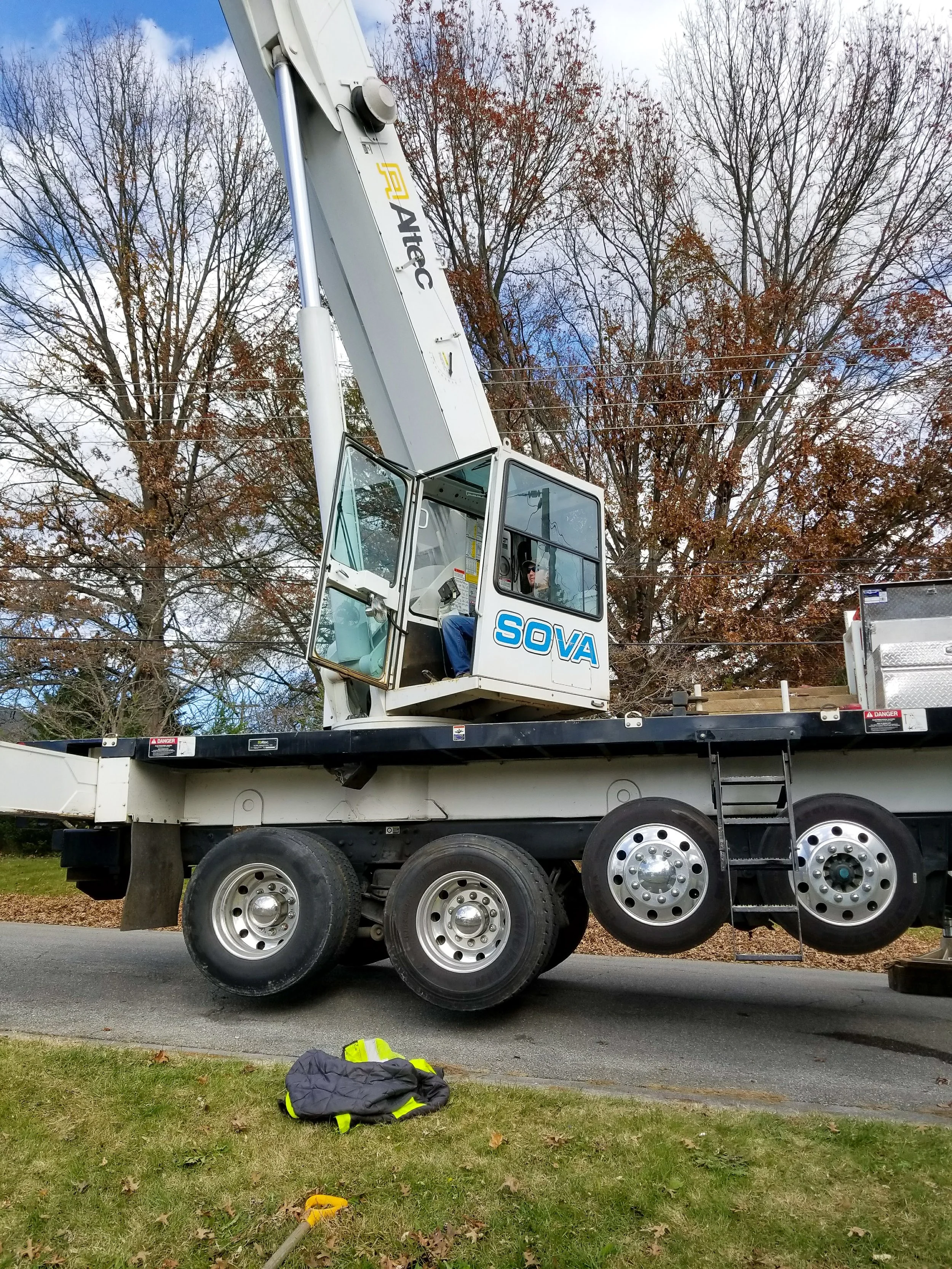A large mobile crane with a white boom and black platform, labeled 'SOVA' and 'Altec,' is extended upward, with a worker in the cab. The crane is parked on a residential street, with trees and a partly cloudy sky in the background. There is a high-vi