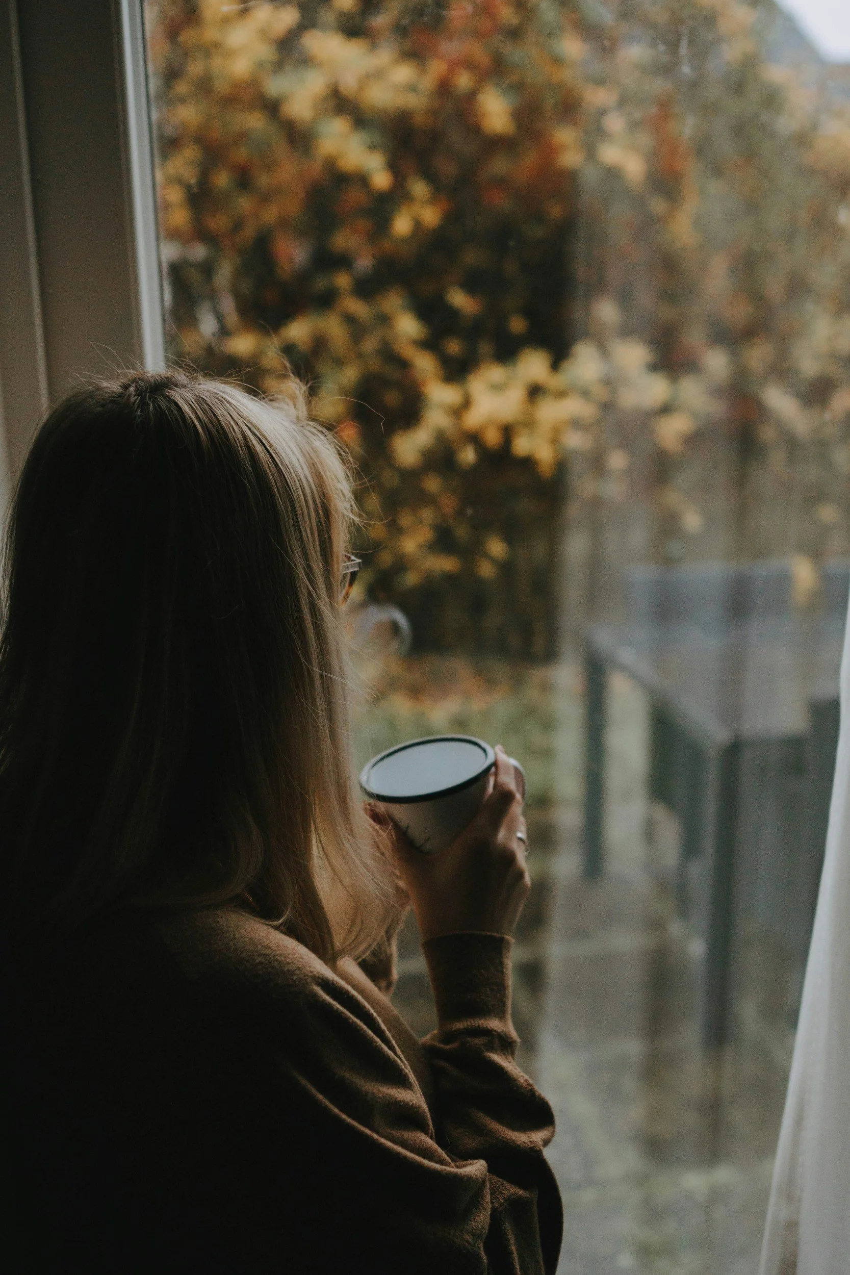 Woman looking out a window holding a mug, representing reflection and self-connection.