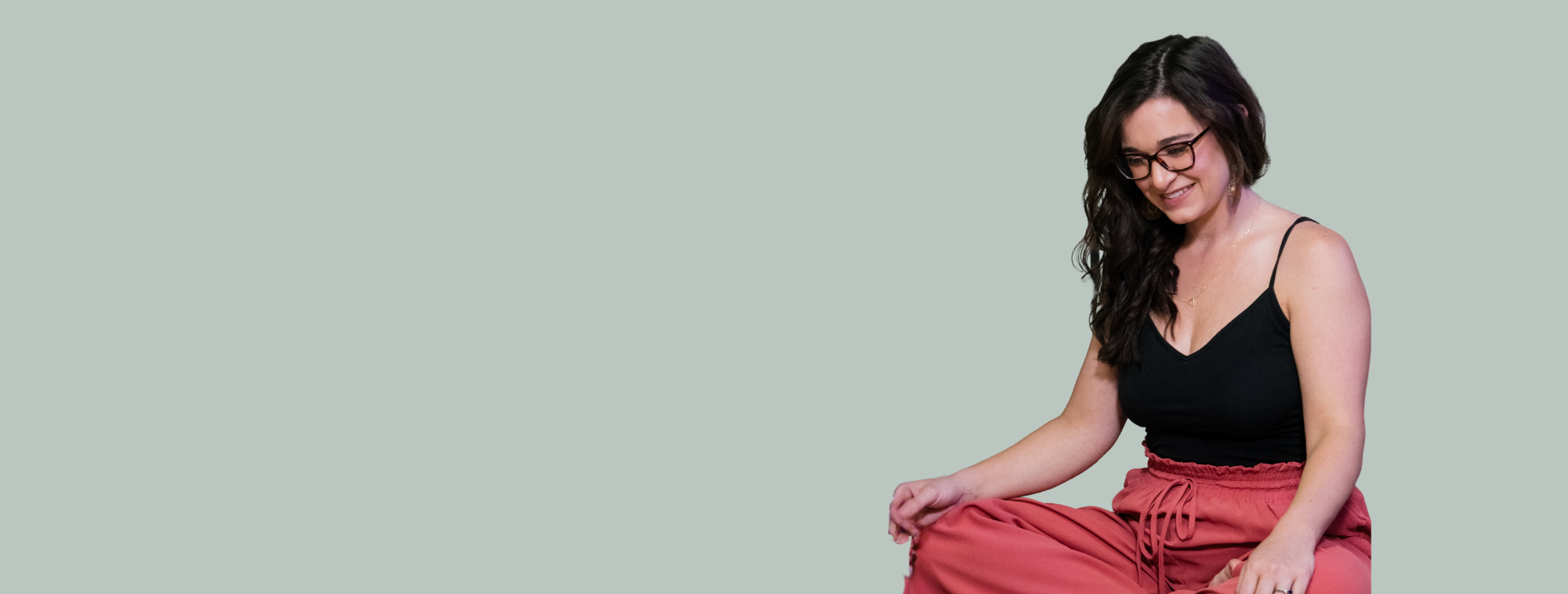 Young woman with glasses, black tank top, and pink pants sitting cross-legged and smiling with her head tilted downward.