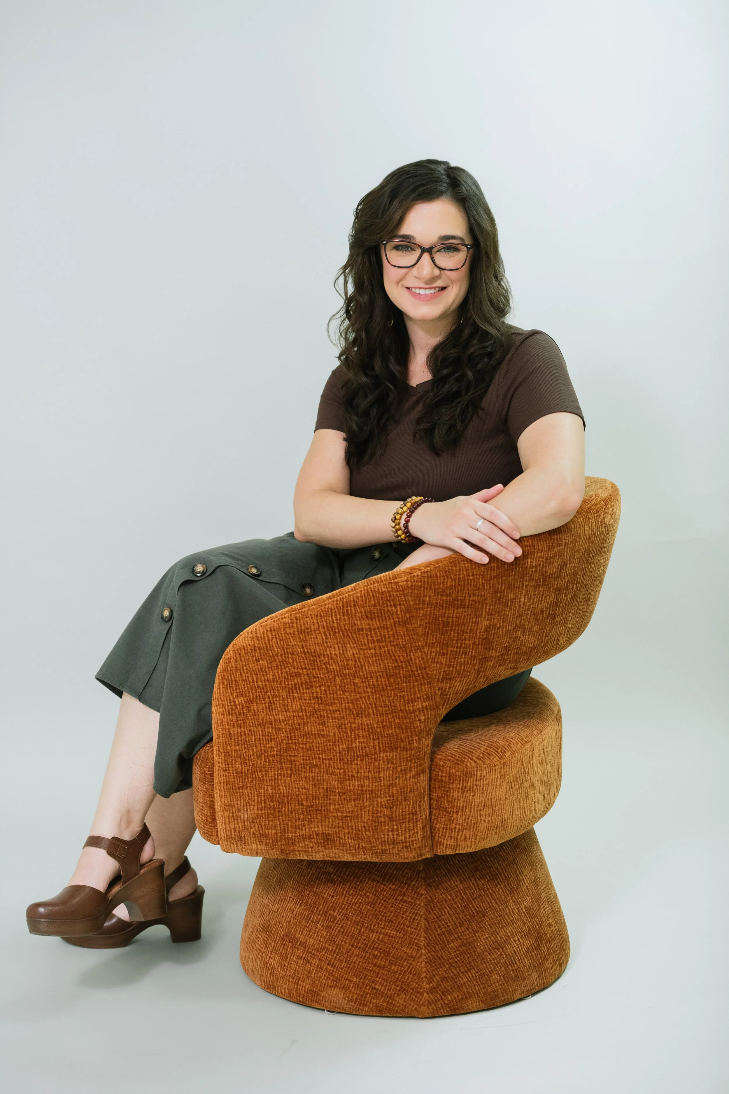 A woman sitting on an orange textured chair, smiling, with glasses, wearing a brown t-shirt, dark green skirt, and brown high heels, against a plain light background.