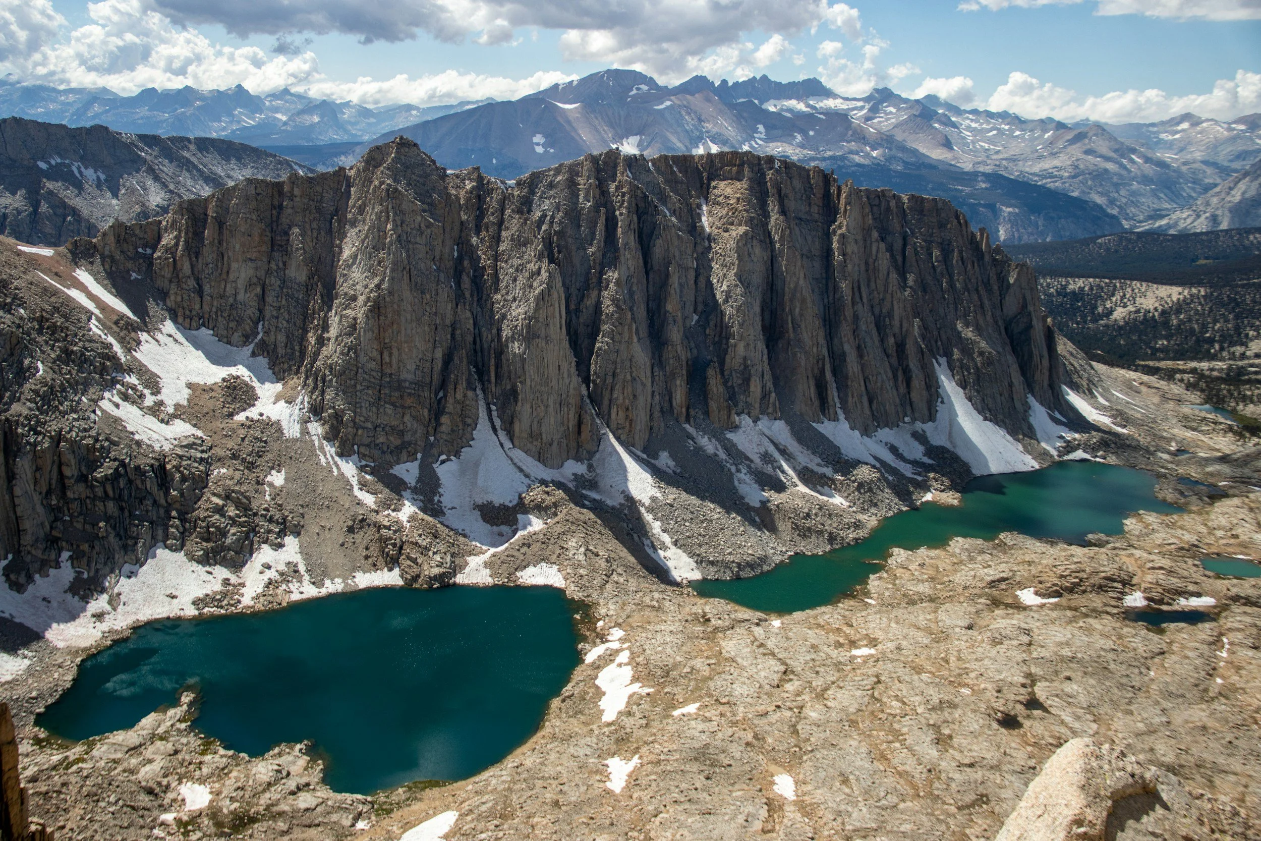 A mountainous landscape with jagged cliffs, snow patches, and two lakes at the base surrounded by rocky terrain, under a partly cloudy sky.