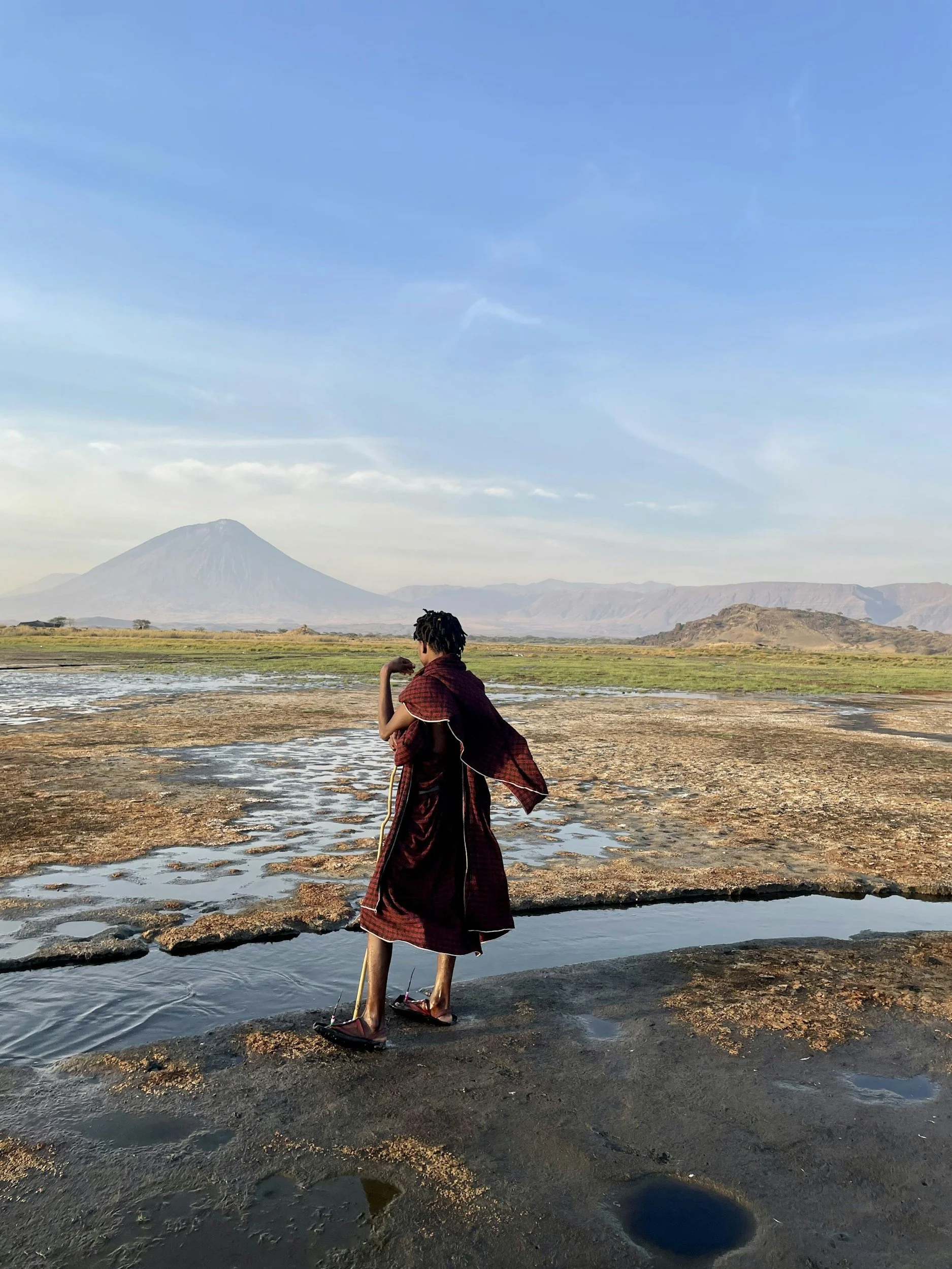A young person in traditional Maasai clothing walking on a rocky terrain with water puddles, with Mount Kilimanjaro and a vast landscape in the background.