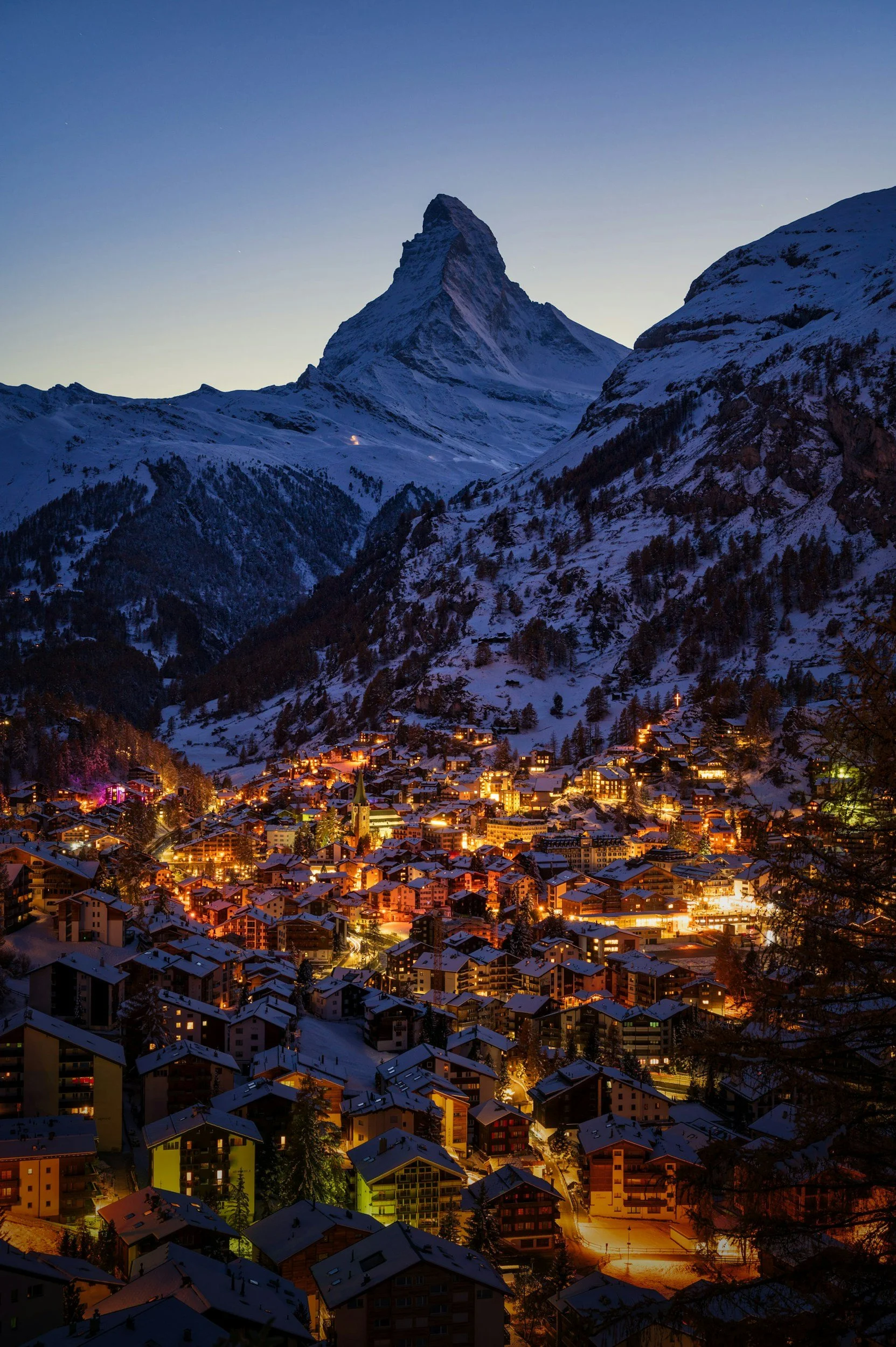 A snow-covered mountain and a town with illuminated buildings at dusk.