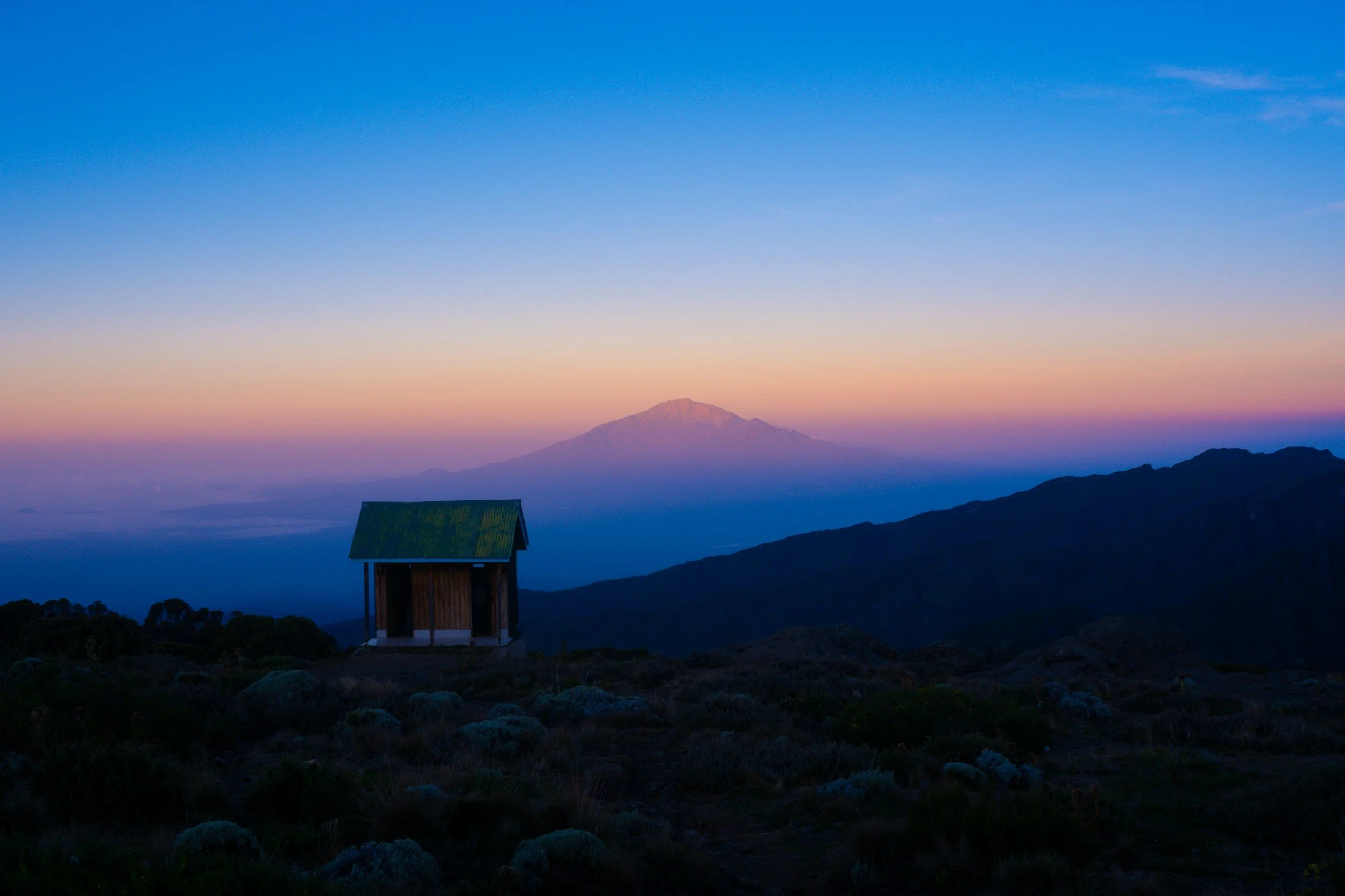 Scenic view of a mountain at sunset with a small wooden shelter in the foreground and a distant snow-capped volcano under a colorful sky.