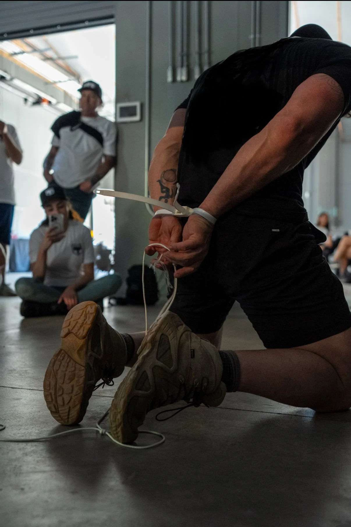 A man kneeling on the floor with hands cuffed behind his back, surrounded by onlookers in an indoor space.