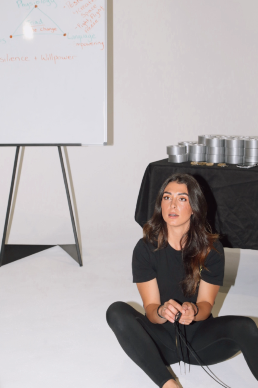 A young woman with long brown hair sitting on the floor in front of a whiteboard and a black table with silver cans on it, in a room with plain white walls.