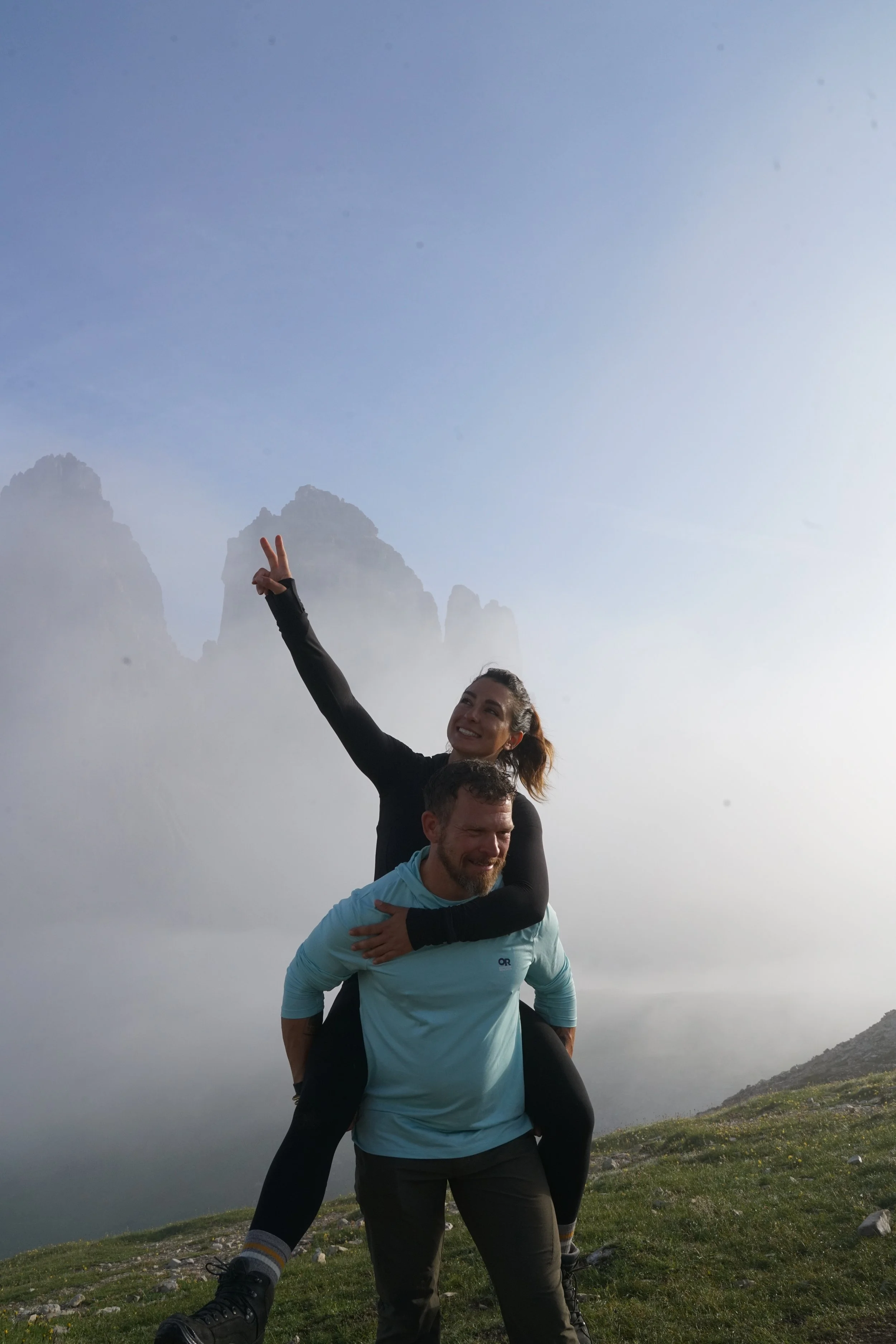 A woman sitting on a man's shoulders, making a peace sign with her hand, in a foggy mountainous landscape.