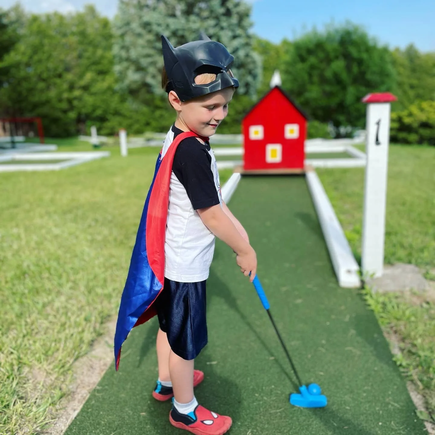 A young boy dressed as a superhero with a cape and mask is playing mini golf outdoors on a green course, holding a blue golf club, with a small red house in the background.