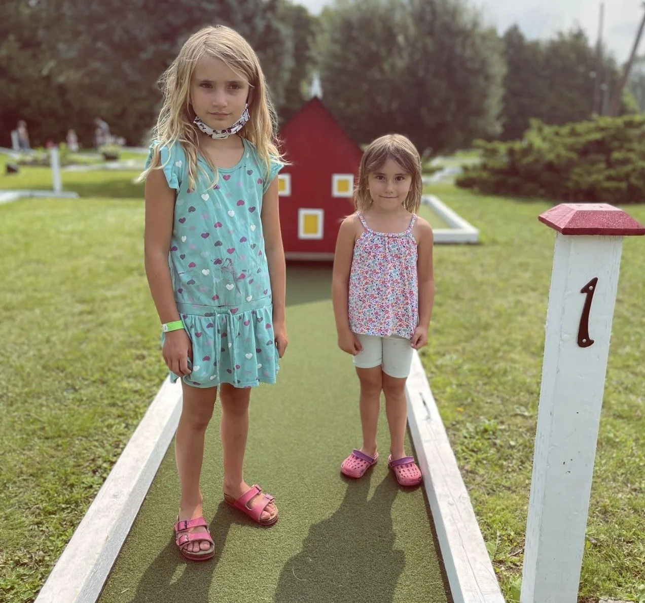 Two young girls standing on a mini-golf hole at an outdoor course during daytime, with greenery and a decorative red house in the background.