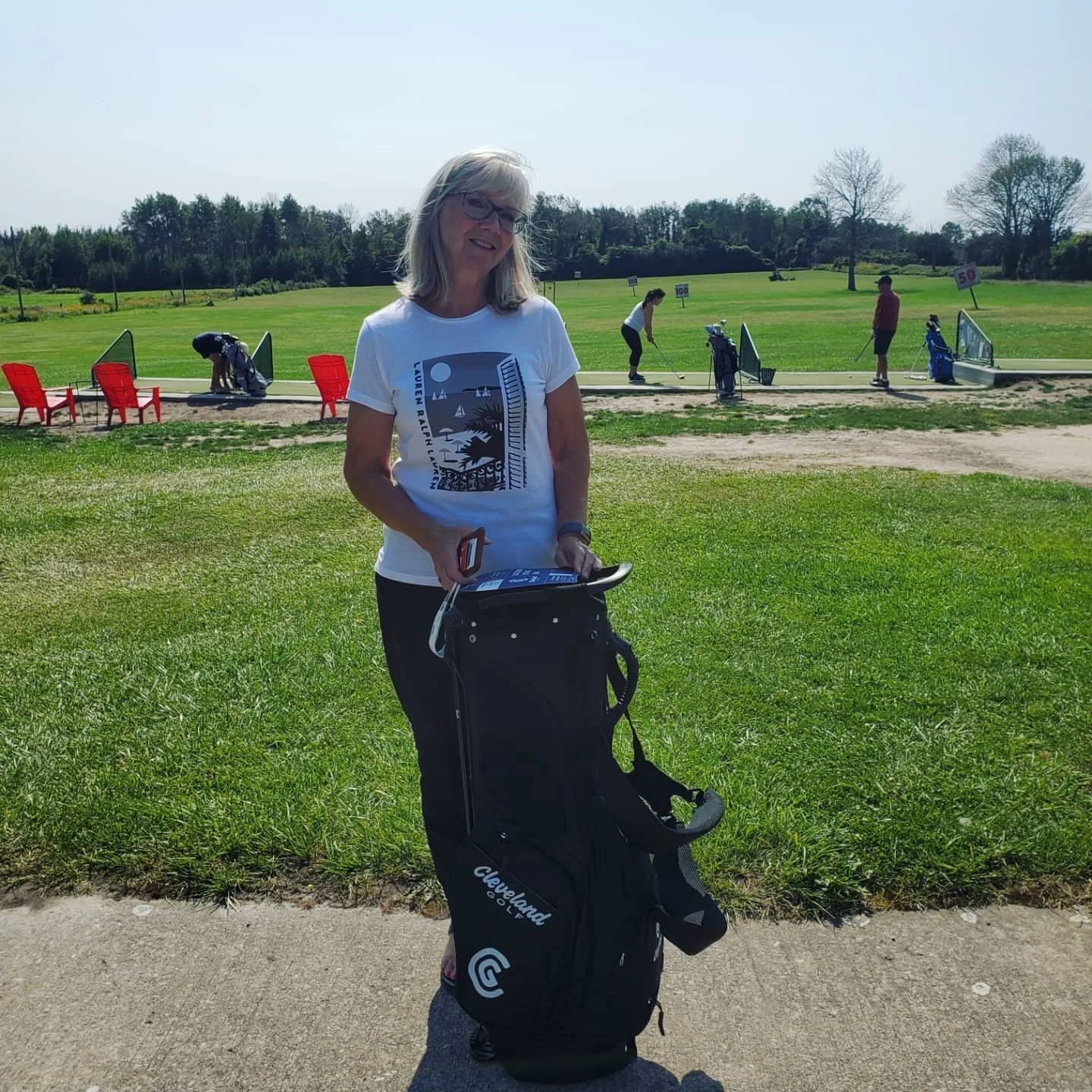 Woman smiling on a golf course holding a golf club and standing next to a golf bag, with a driving range behind her where three people are practicing and red chairs are lined up.