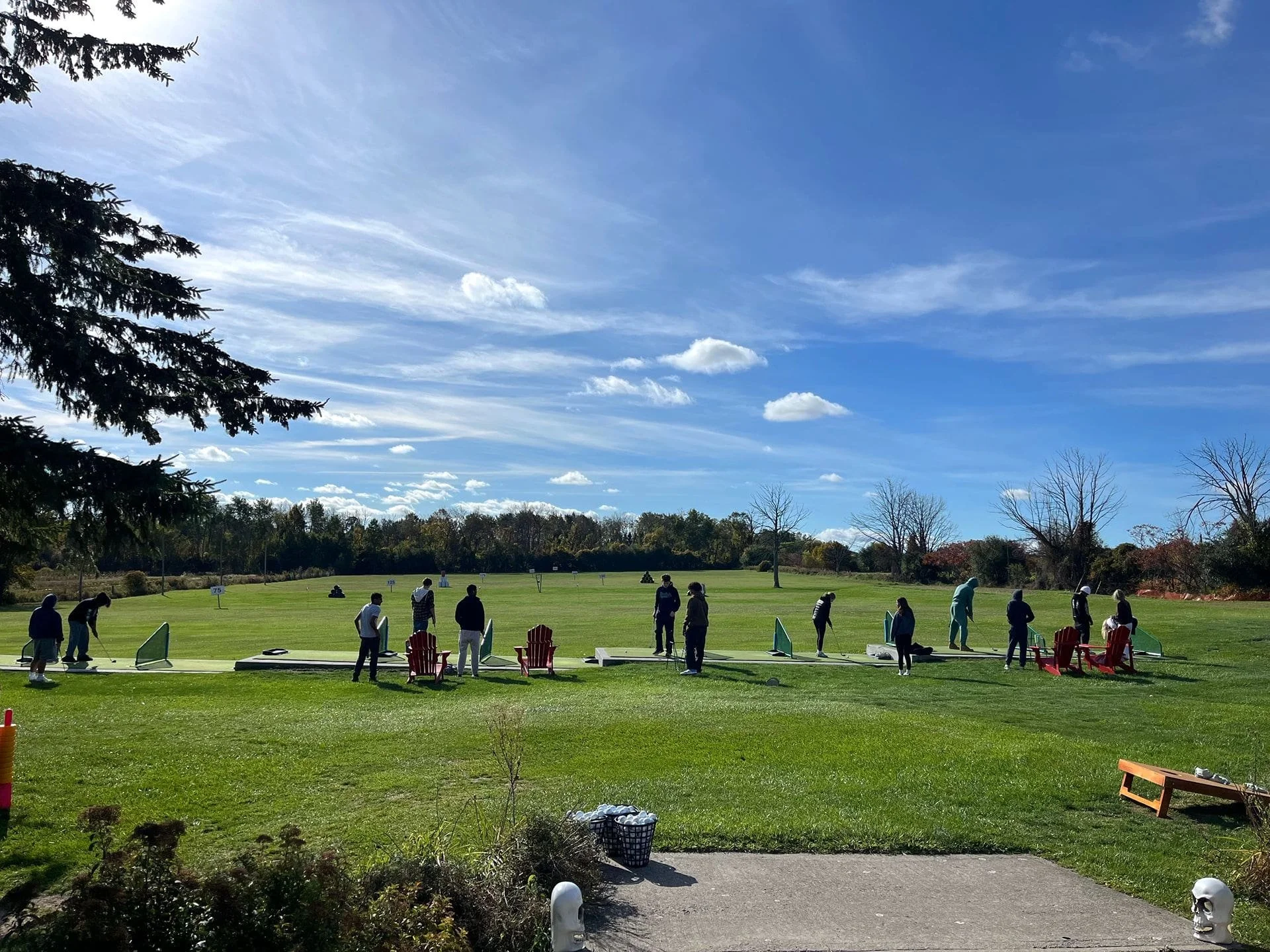 People practicing on the driving range outdoors on a sunny day with a clear blue sky and scattered clouds.
