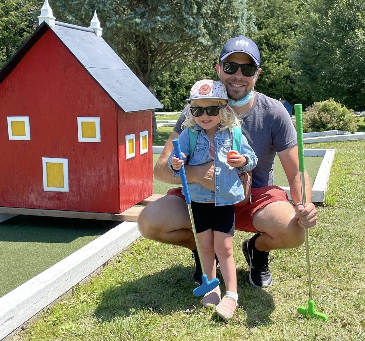 A man and a young girl with sunglasses, hats, and casual clothing, holding mini golf clubs, kneeling and smiling next to a small red house prop on a mini golf course outdoors on a sunny day.
