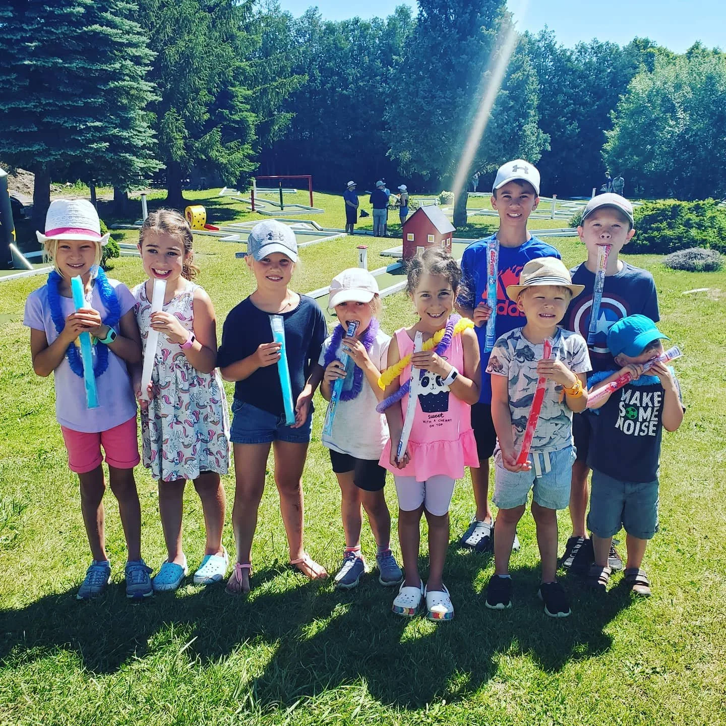 Group of ten children standing outdoors on a mini putt course, holding freezies, with a sunny background and trees.