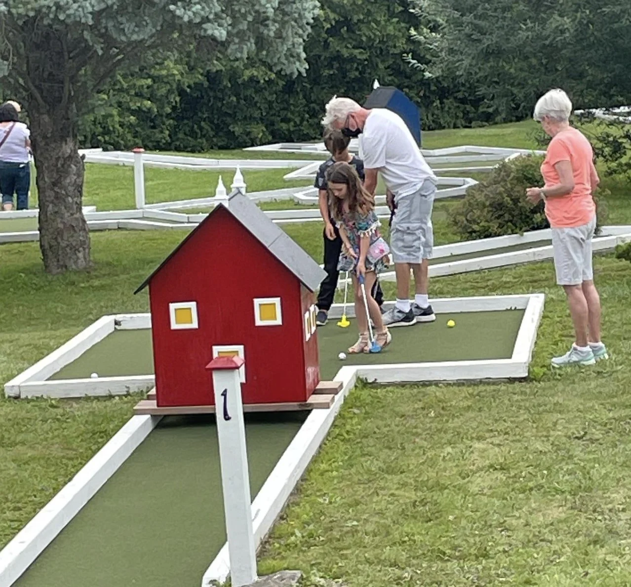 People playing mini golf outdoors on a course with green artificial turf and themed obstacles, including a red house structure, surrounded by trees and white fencing.