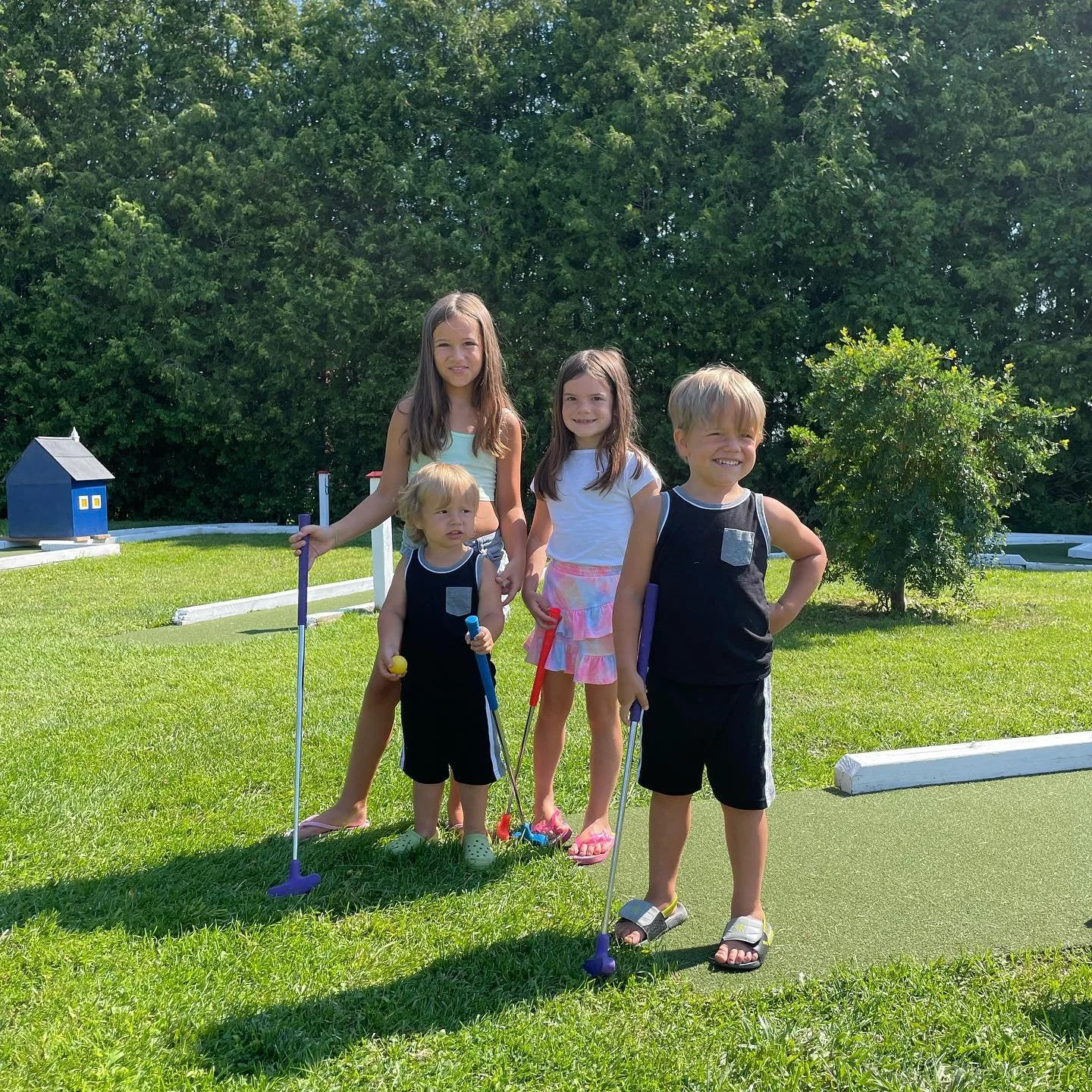 Four children playing mini golf outdoors on a sunny day, standing on a green course with trees and bushes in the background.