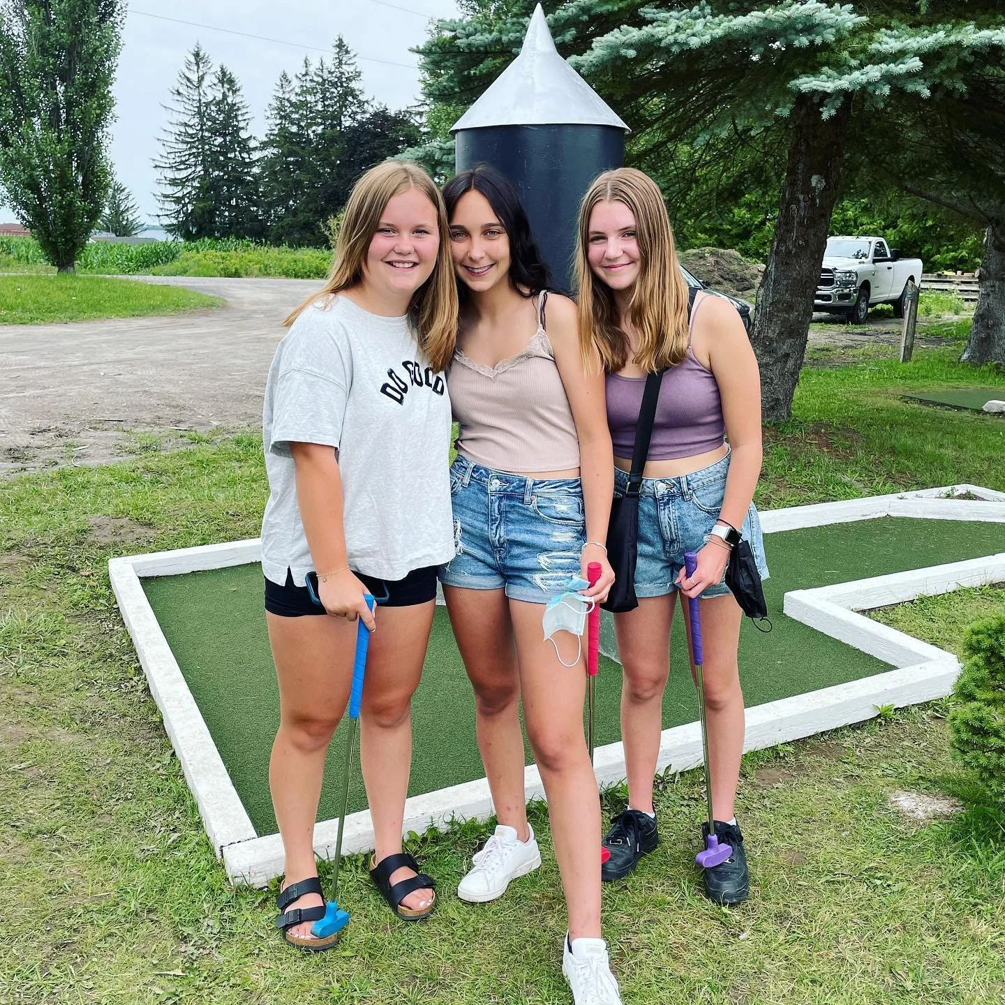 Three young women standing on mini golf course. They are smiling, holding mini golf clubs, and wearing casual summer clothes.