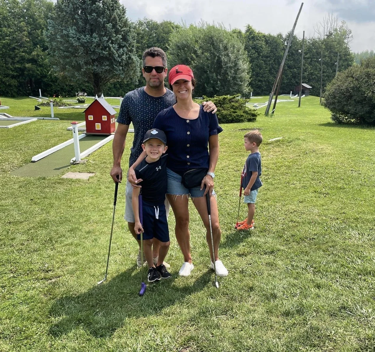 A family of four with two young boys standing on a golf course, smiling at the camera, with one boy holding a golf club.