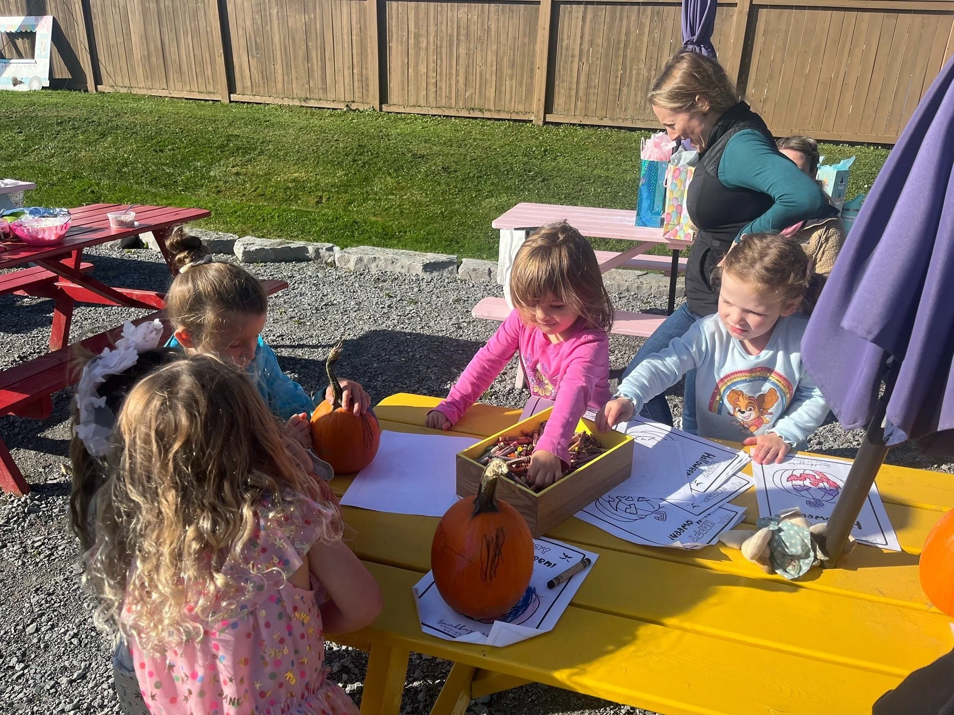 Children gathered around a yellow table outside, doing arts and crafts with pumpkins. An adult supervises. There are coloring pages, crayons, and pumpkins on the table. The scene is sunny with a wooden fence and grassy area in the background.