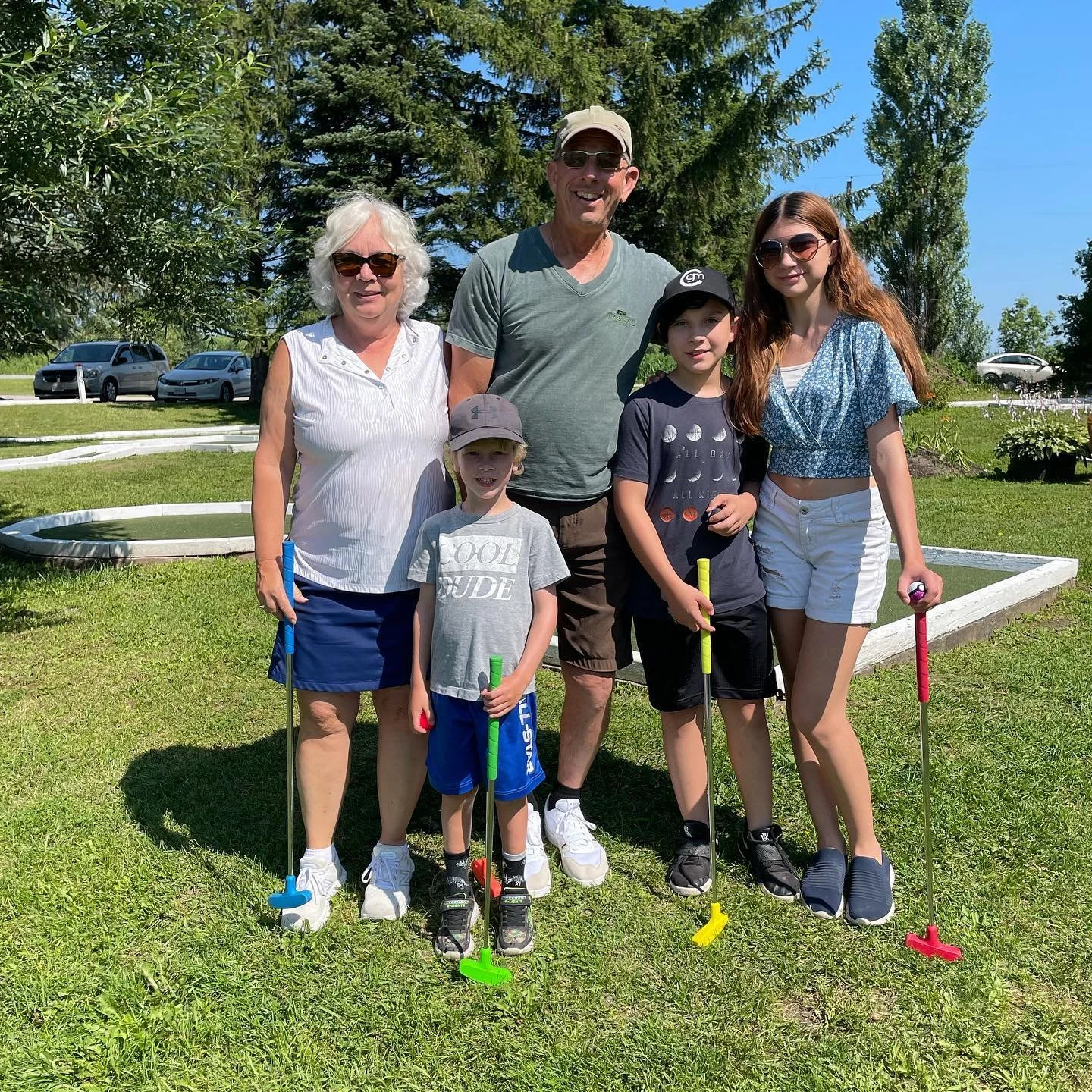 A family of six standing on a lawn with mini golf putters, smiling, with trees, and a clear blue sky in the background.