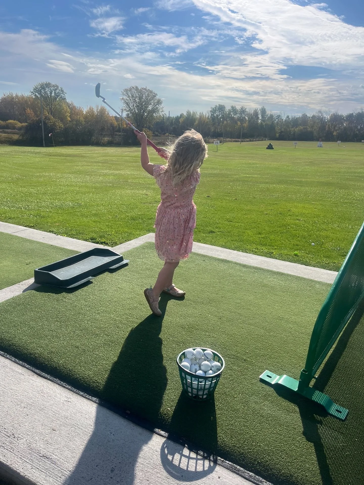 Young girl in pink dress practicing golf at driving range with basket of golf balls nearby and a golf club in her hands