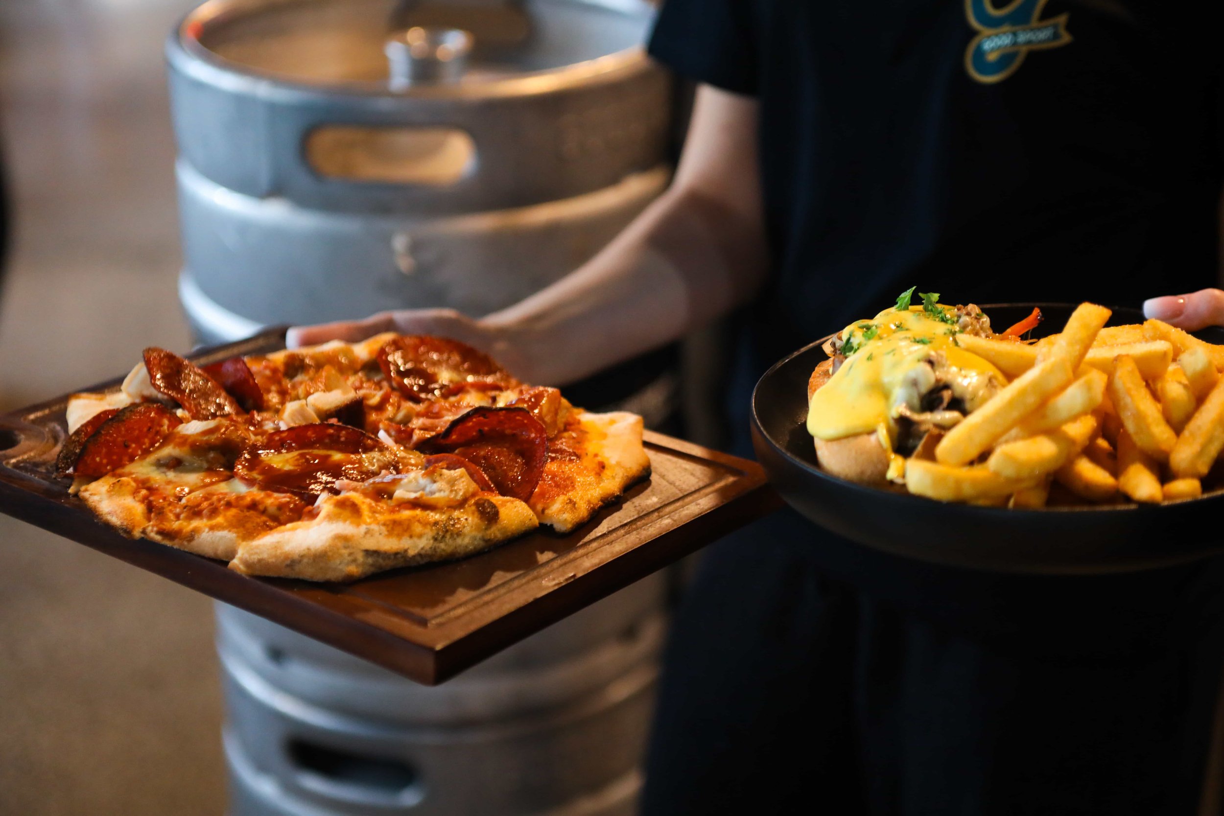 Person holding a wooden tray with a slice of pepperoni pizza, and a plate of loaded fries topped with cheese and greens at Good Sport Napier