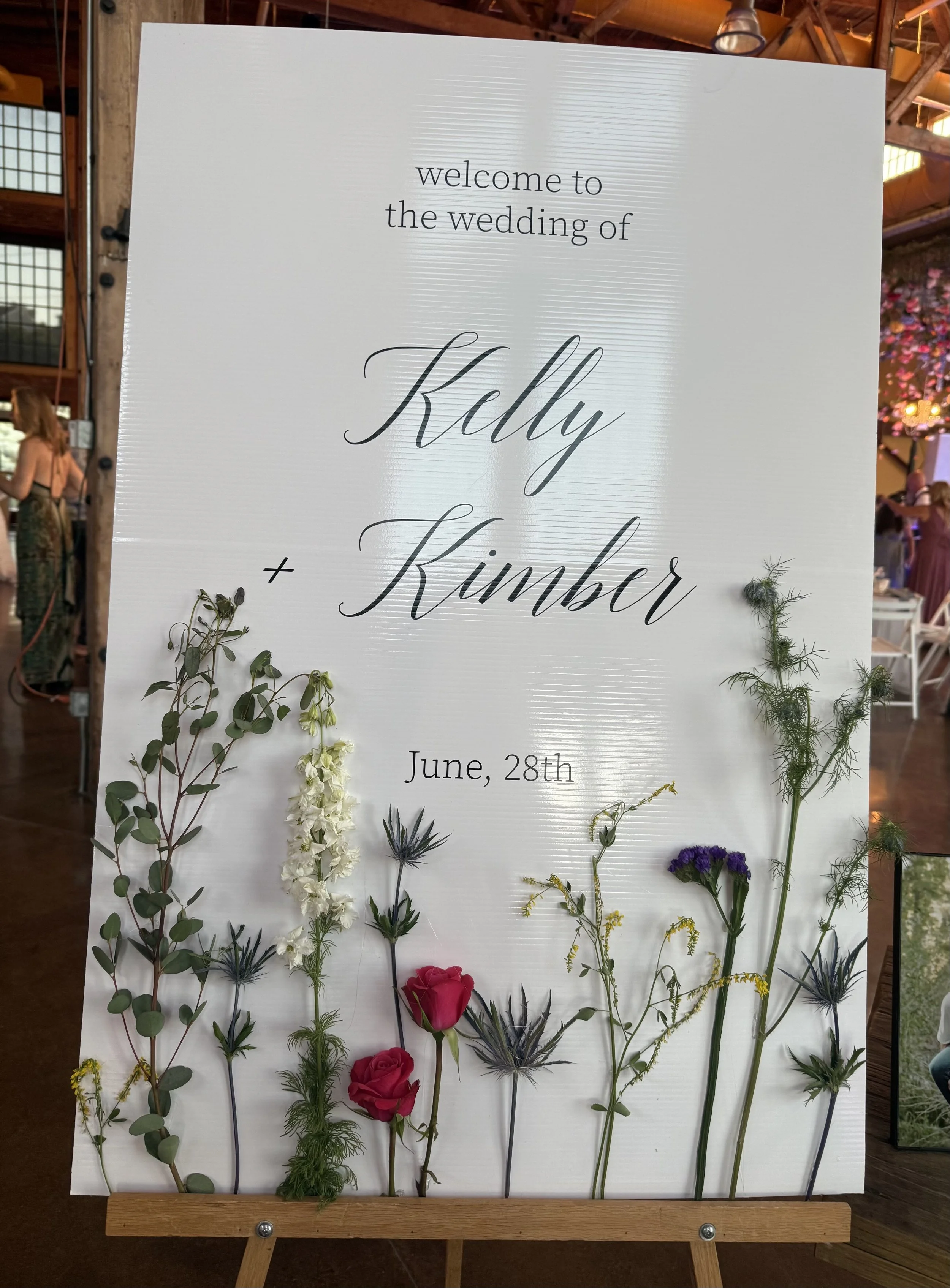 Wedding welcome sign with the names Kelly and Kumber, dated June 28th. Decorated with various wildflowers at the bottom.