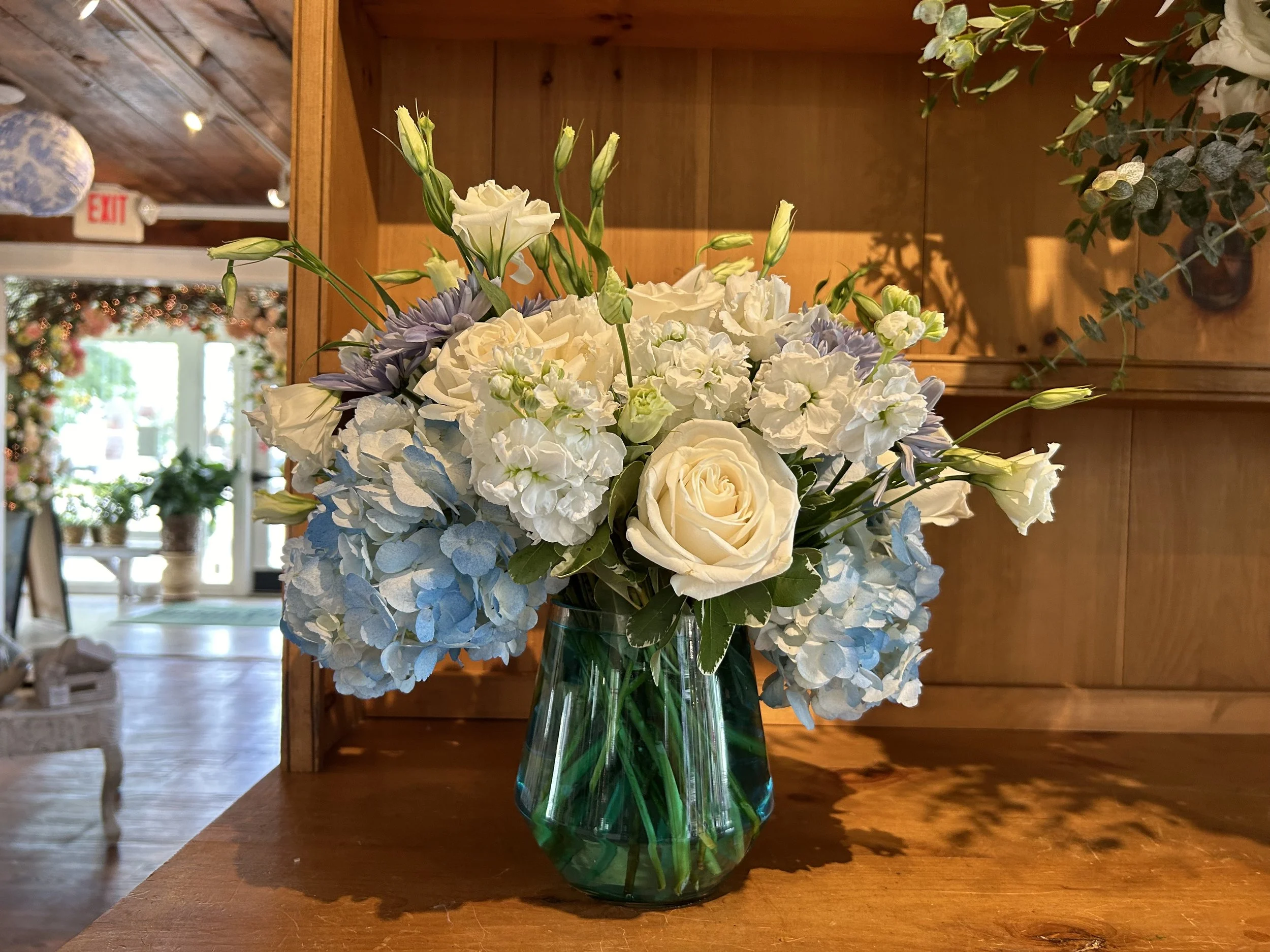A flower arrangement with white roses, blue hydrangeas, and other white flowers in a green glass vase placed on a wooden surface inside a room.