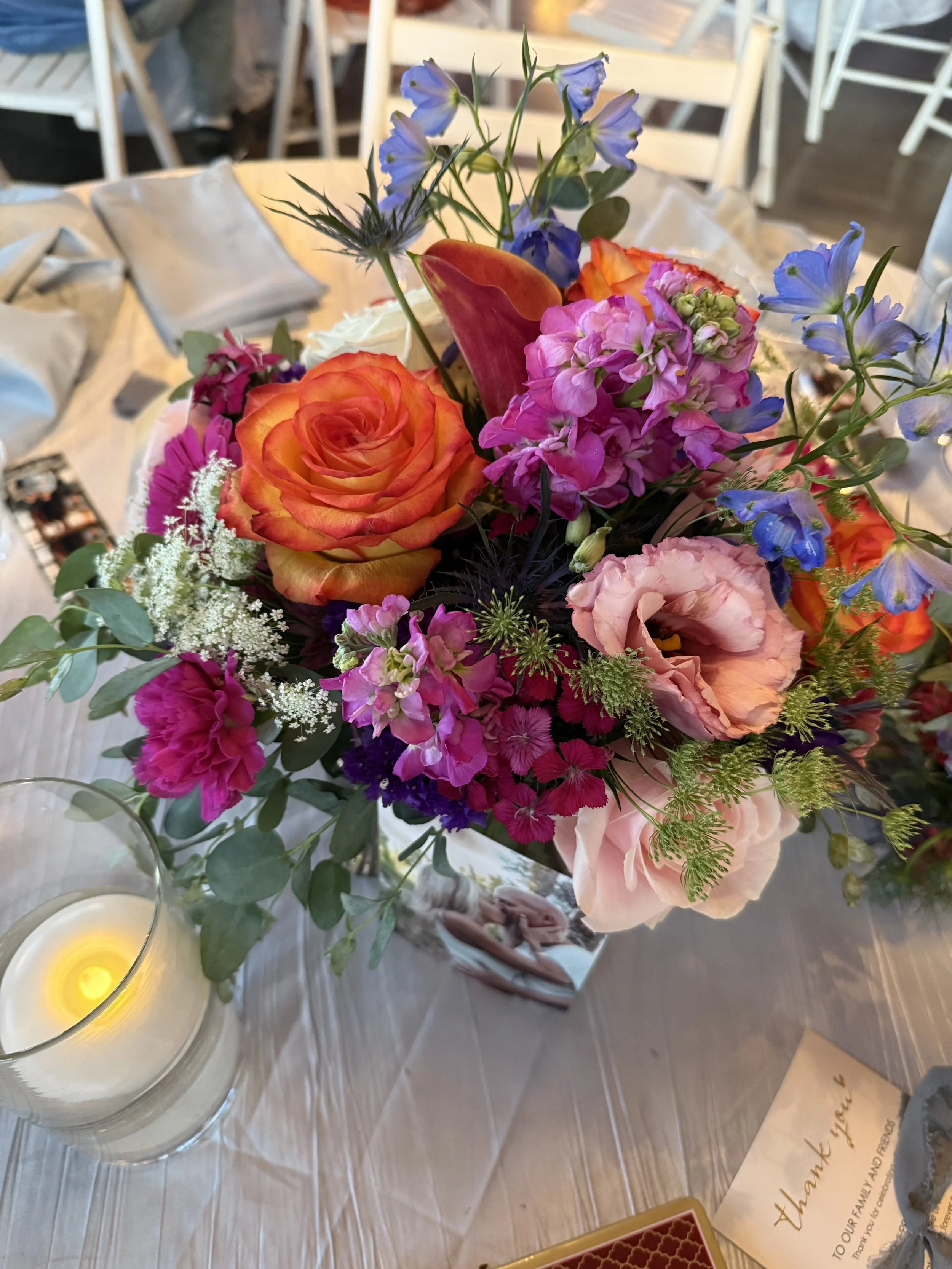 A colorful flower bouquet with roses, pink, purple, and blue flowers on a table with a candle, a photo, and a thank you card.