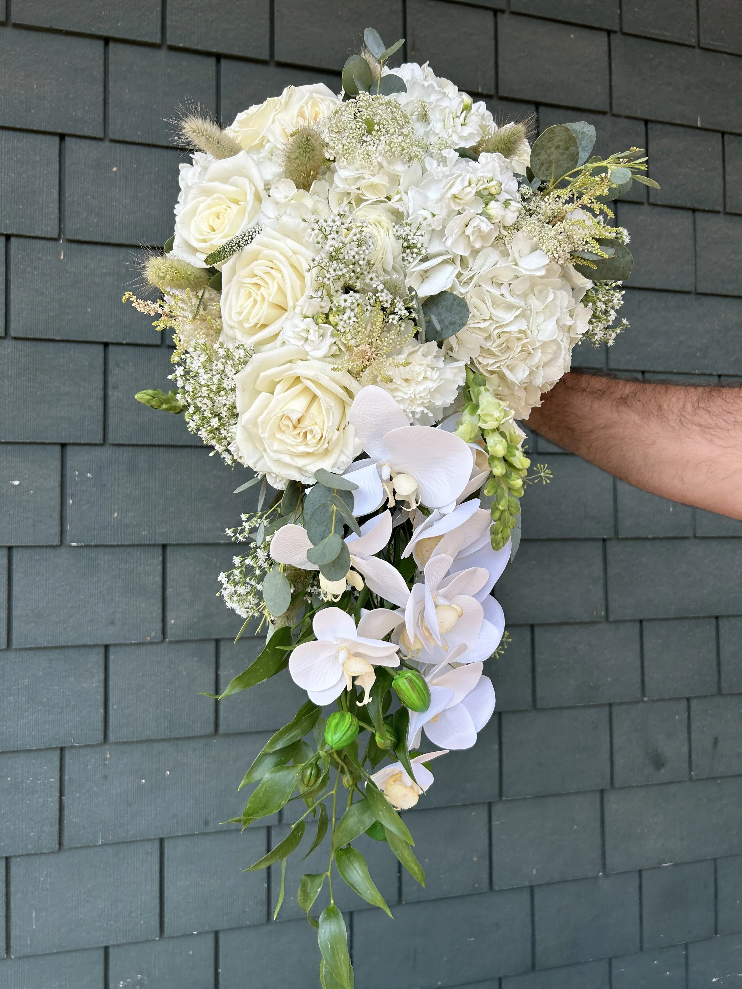 A cascading wedding bouquet featuring white roses, hydrangeas, orchids, and greenery, held by a person against a dark green tiled wall.