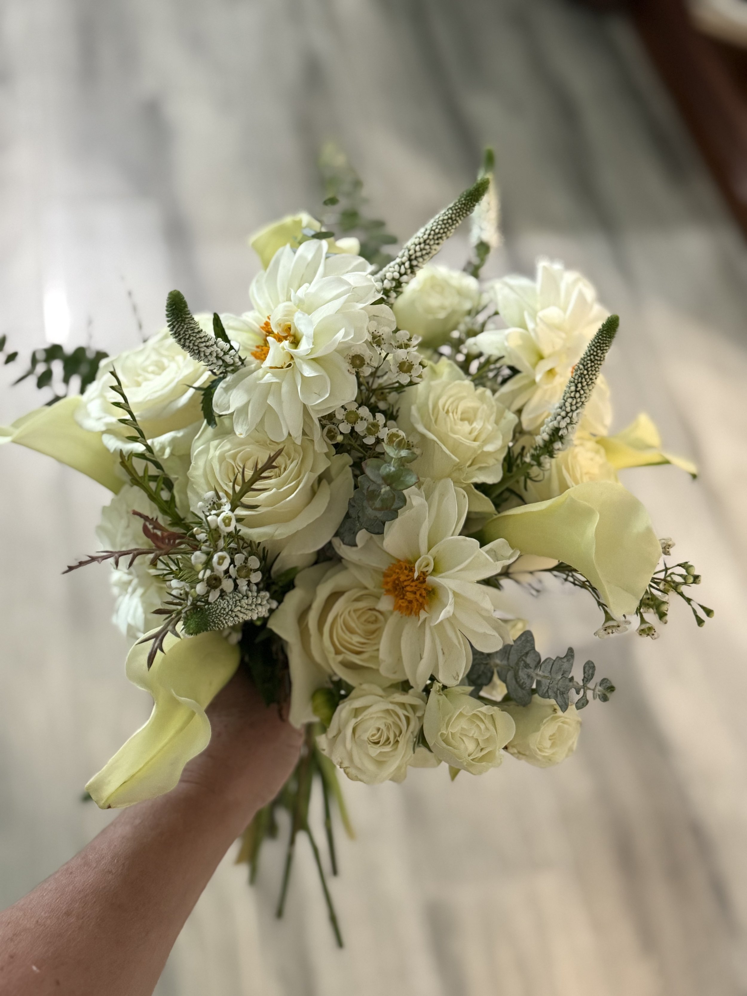 A hand holding a bouquet of white flowers including roses, dahlias, calla lilies, and other greenery on a wooden floor