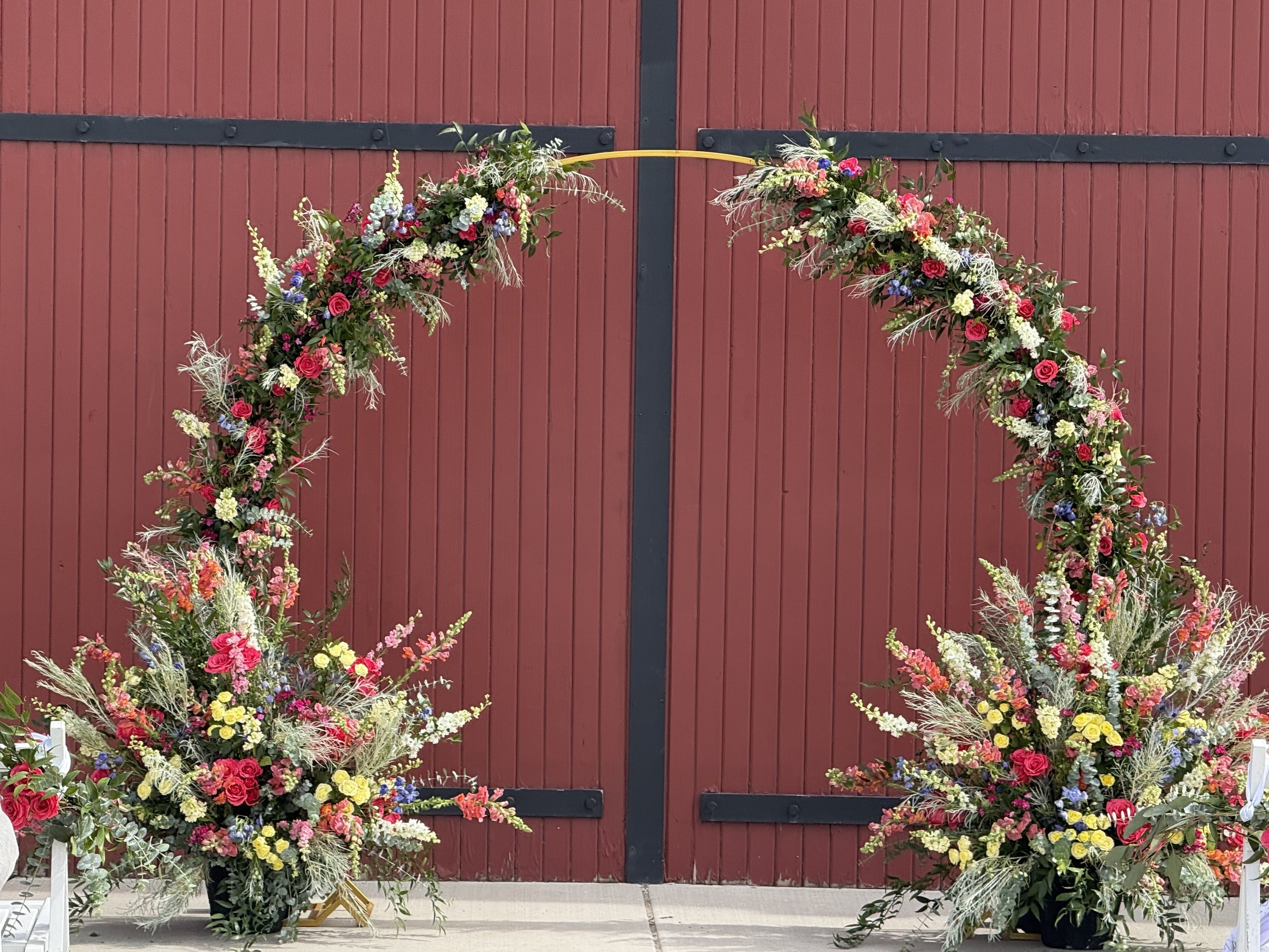Floral wedding arch with pink, yellow, white, purple, and green flowers in front of a red barn door.