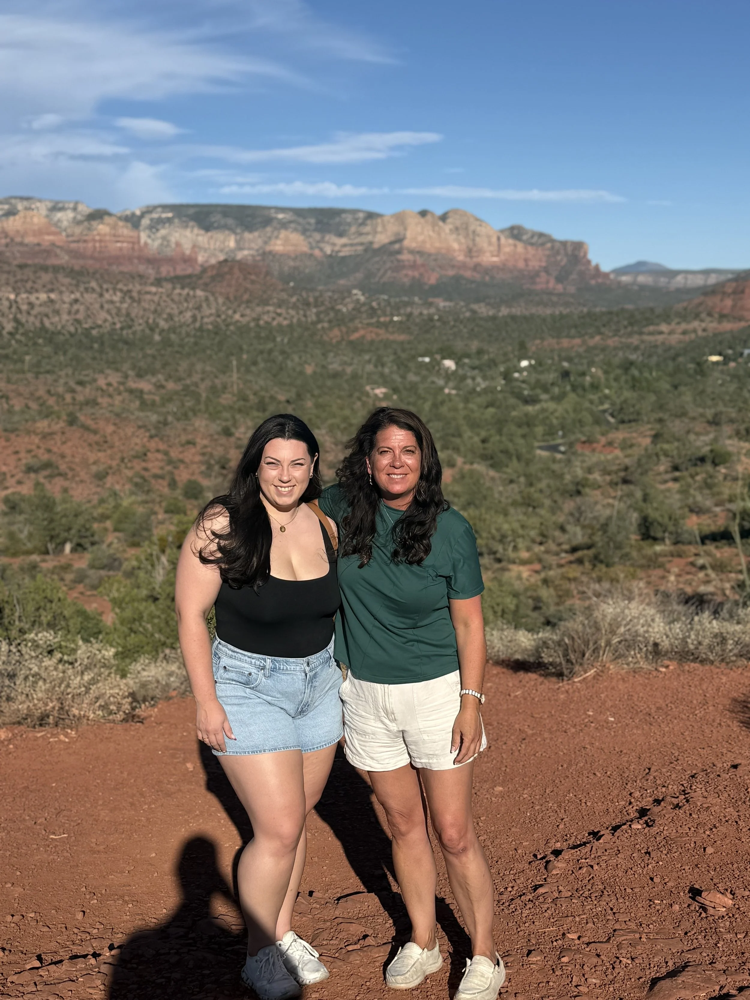 Two women smiling and standing on a dirt trail with red rocks and desert vegetation, with a mountain range and blue sky in the background.