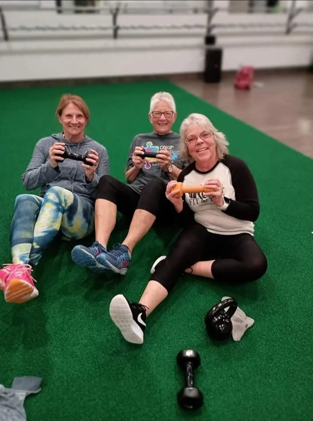 Three women sitting on green turf, smiling and holding small weights and a gaming controller in a fitness or recreation facility.