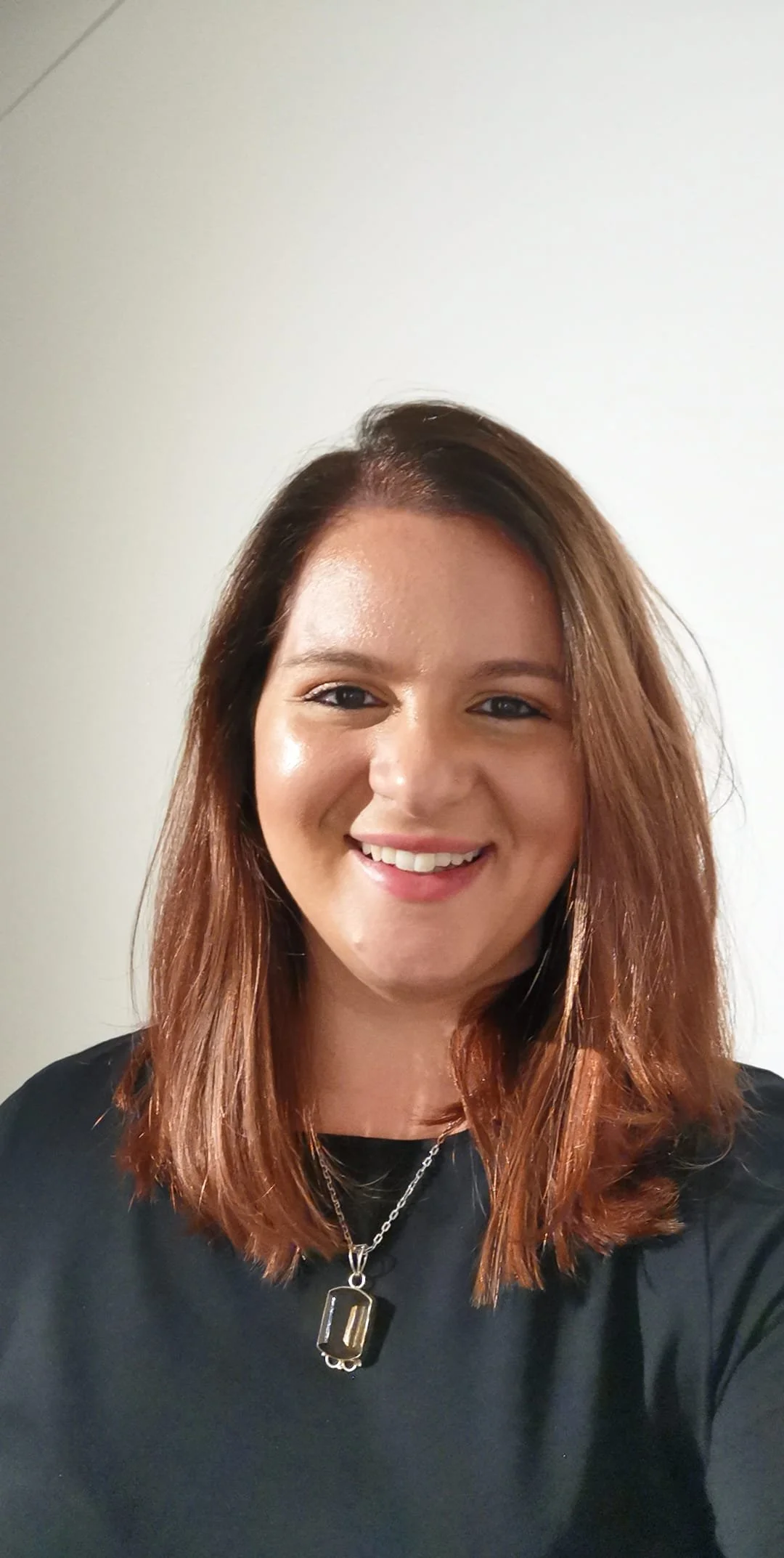 A smiling woman with shoulder-length reddish-brown hair, wearing a black shirt and layered silver necklaces, standing against a plain white wall.