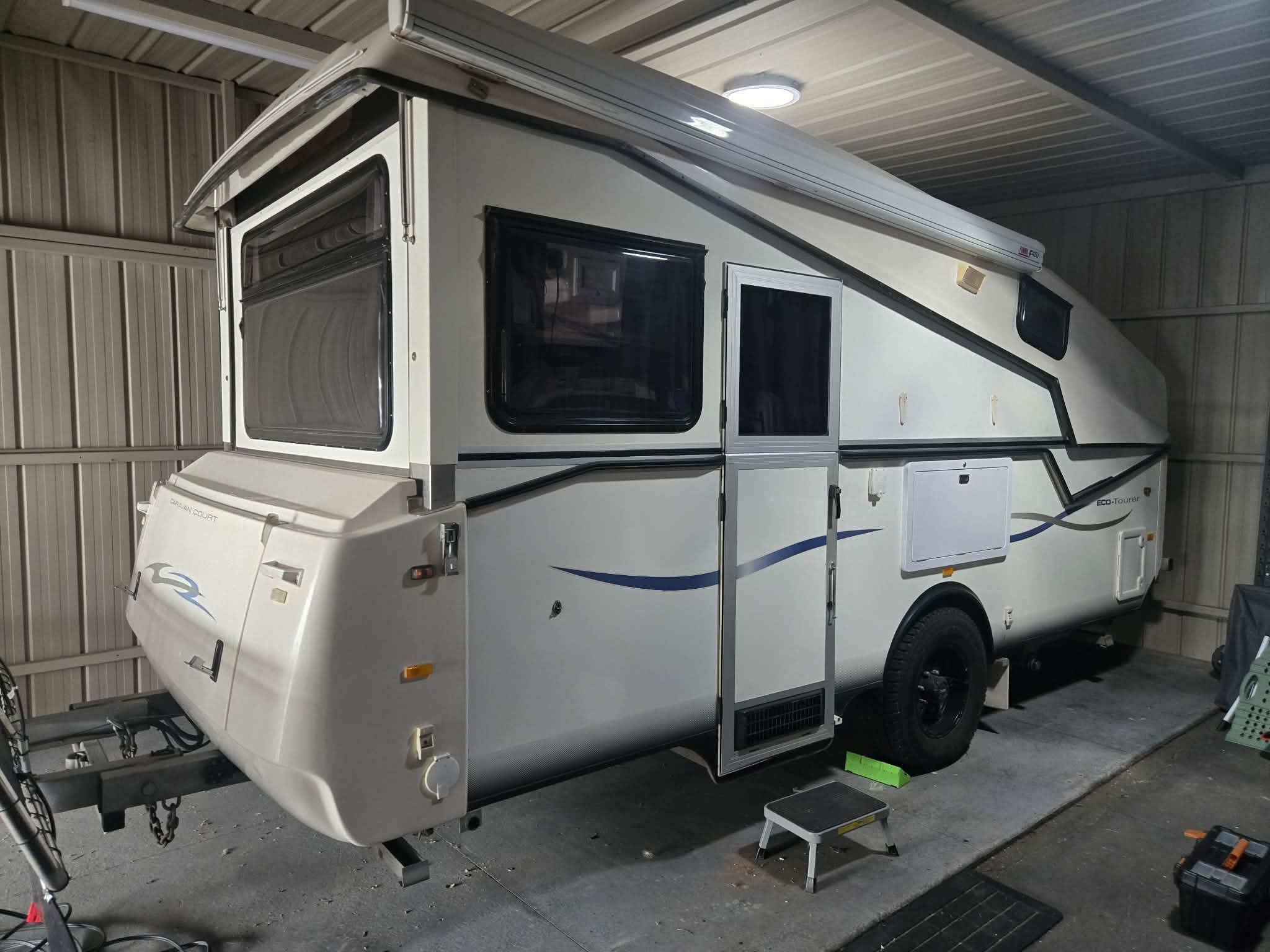 A white travel trailer with black and blue accents parked inside a storage shed, with a step stool and various items around it.