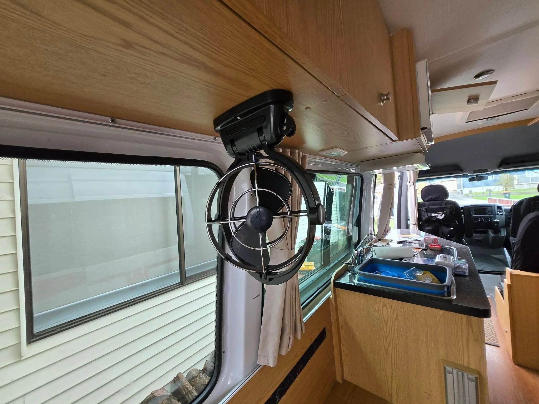 Inside a motorhome or RV with a small fan mounted on the ceiling above the kitchen counter, which has a sink, dishes, and various items on it. The interior features wood cabinetry and a large window with curtains.