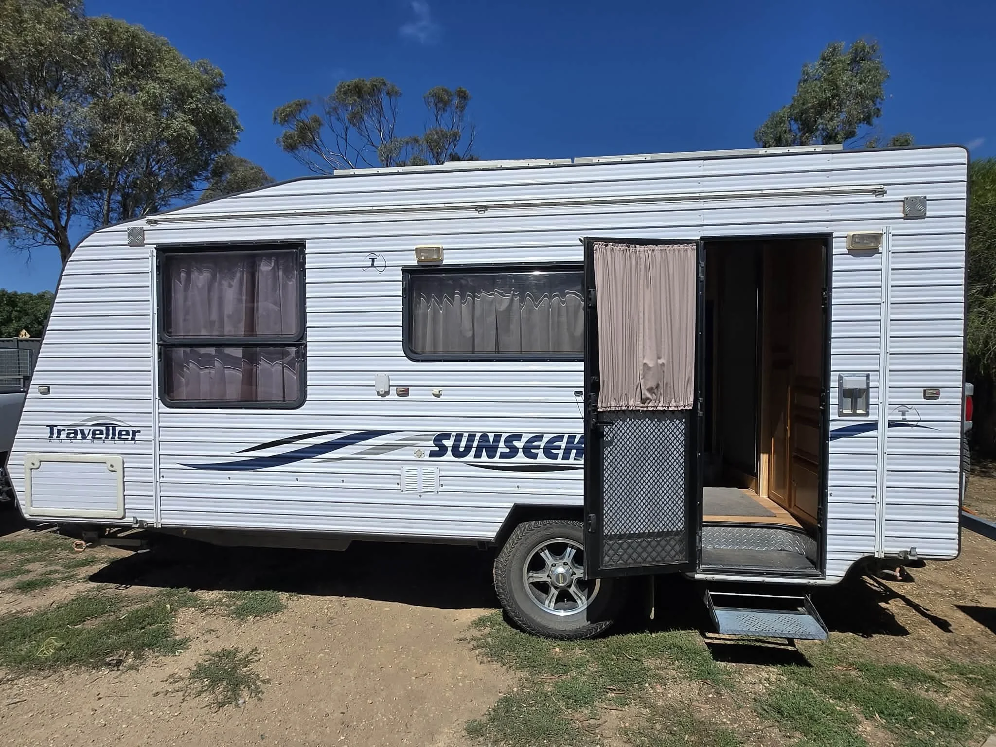 A white travel trailer with opened door, two windows with curtains, and branding that says 'Sunseeker' and 'Traveller' parked on a dirt patch with grass, trees, and a blue sky in the background.