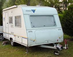 A white travel trailer with black and blue accents parked inside a storage shed, with a step stool and various items around it.