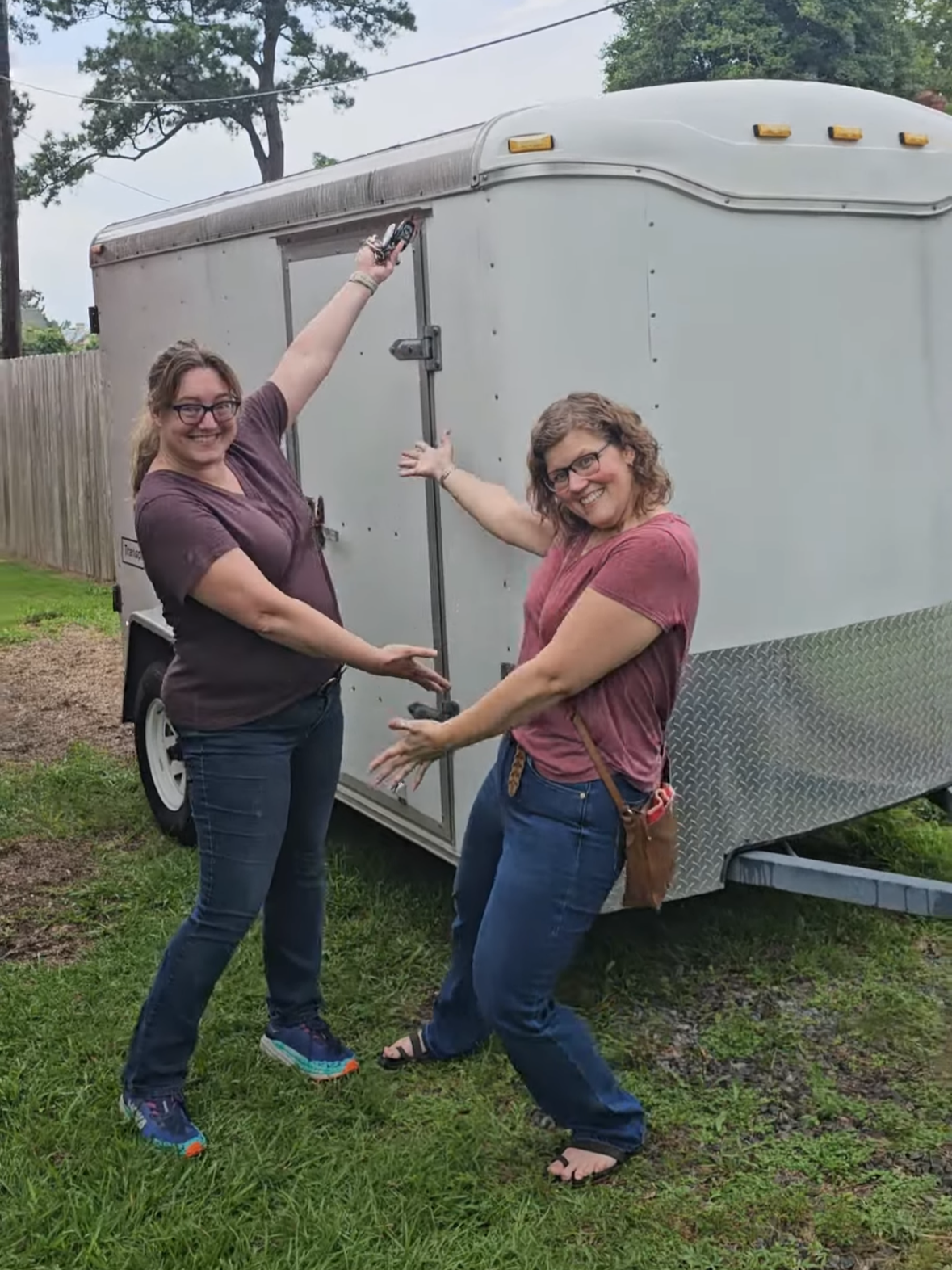 Two women smiling and posing in front of a silver enclosed trailer on grass.