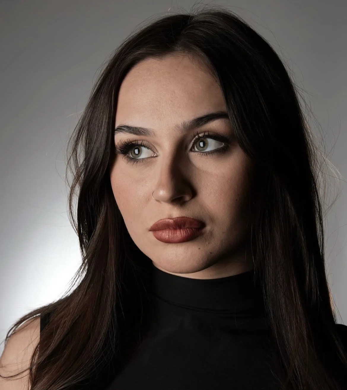 Close-up portrait of a woman with long dark hair, wearing a black sleeveless top, looking away from the camera with a serious expression.