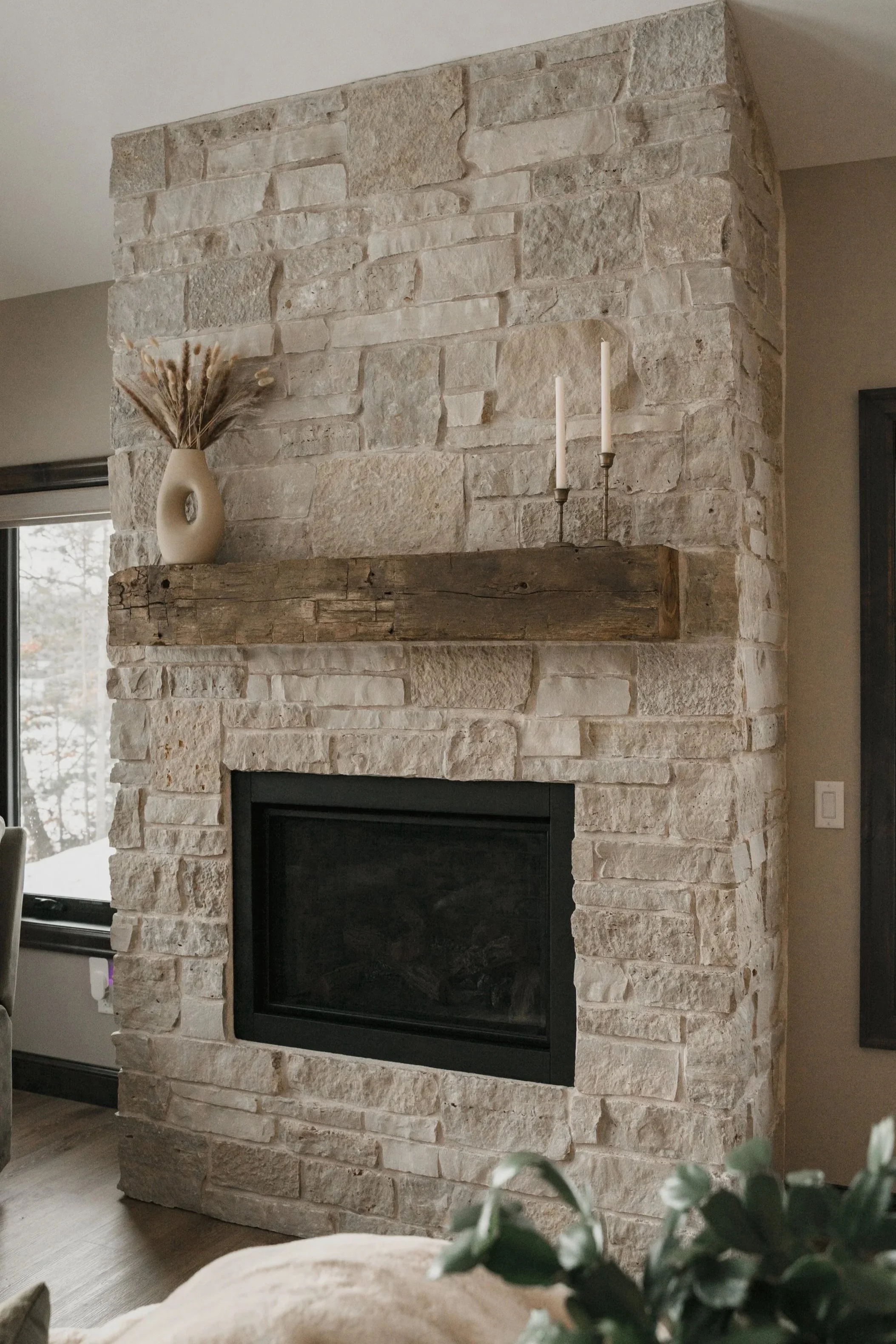 Living room fireplace with a stone facade, a wooden mantel shelf, a white ceramic vase with dried flowers, and two white candles in brass holders.