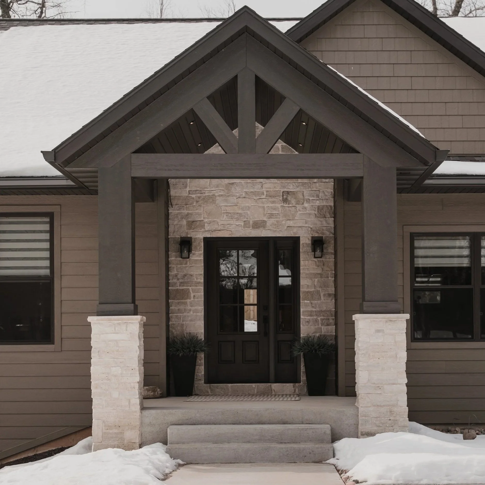 Front entrance of a house with stone columns, a black door, and a gabled roof with dark shingles, snow on the ground and on the roof.