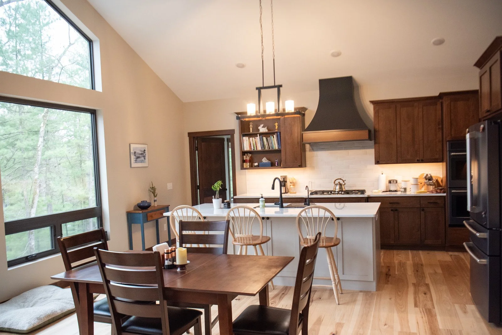 Open-concept kitchen and dining area with wood cabinetry, large island, dining table, and floor-to-ceiling windows overlooking wooded scenery.