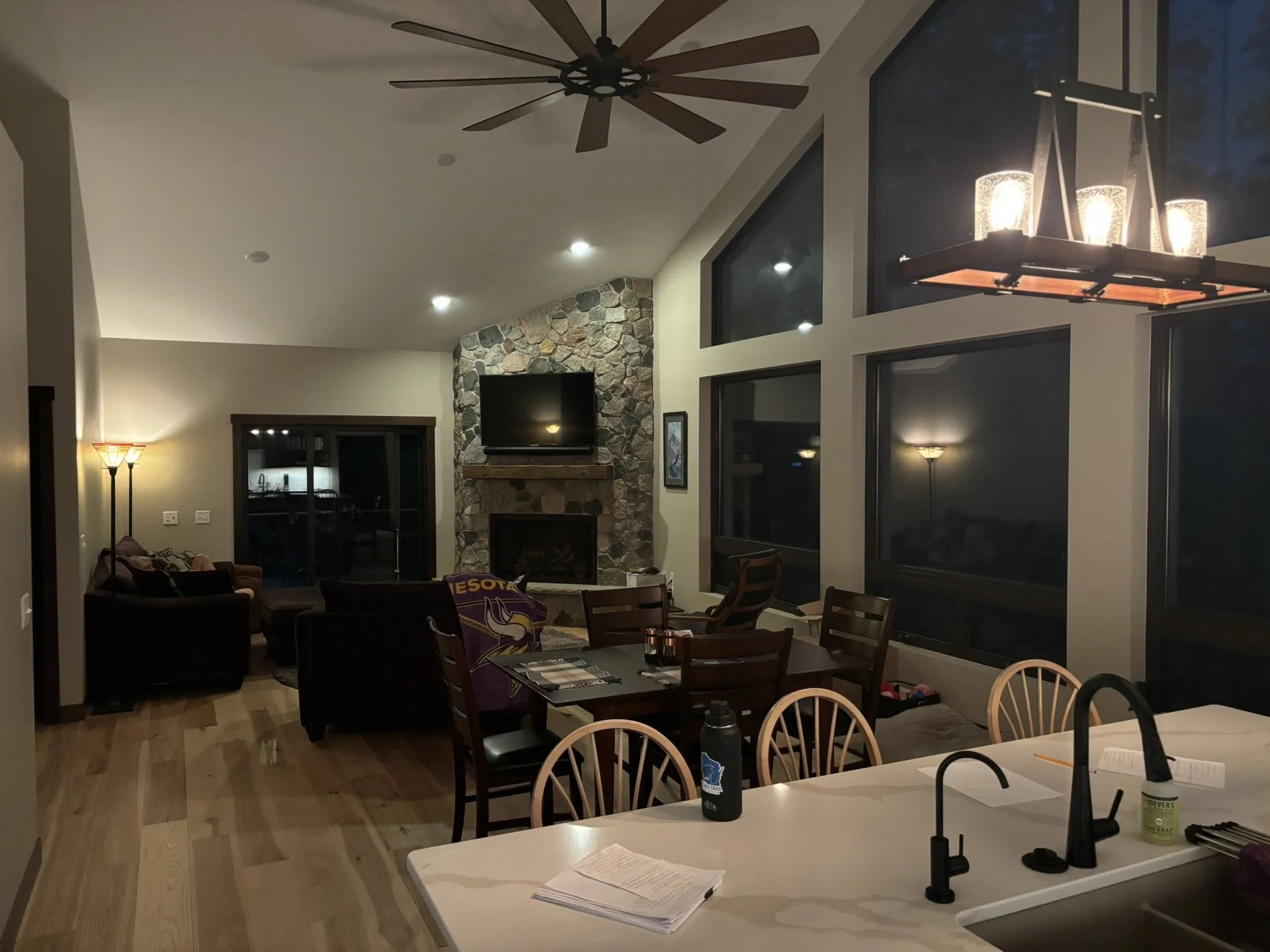 Open-concept kitchen and living room with high ceilings, large windows, stone fireplace, and warm interior lighting.