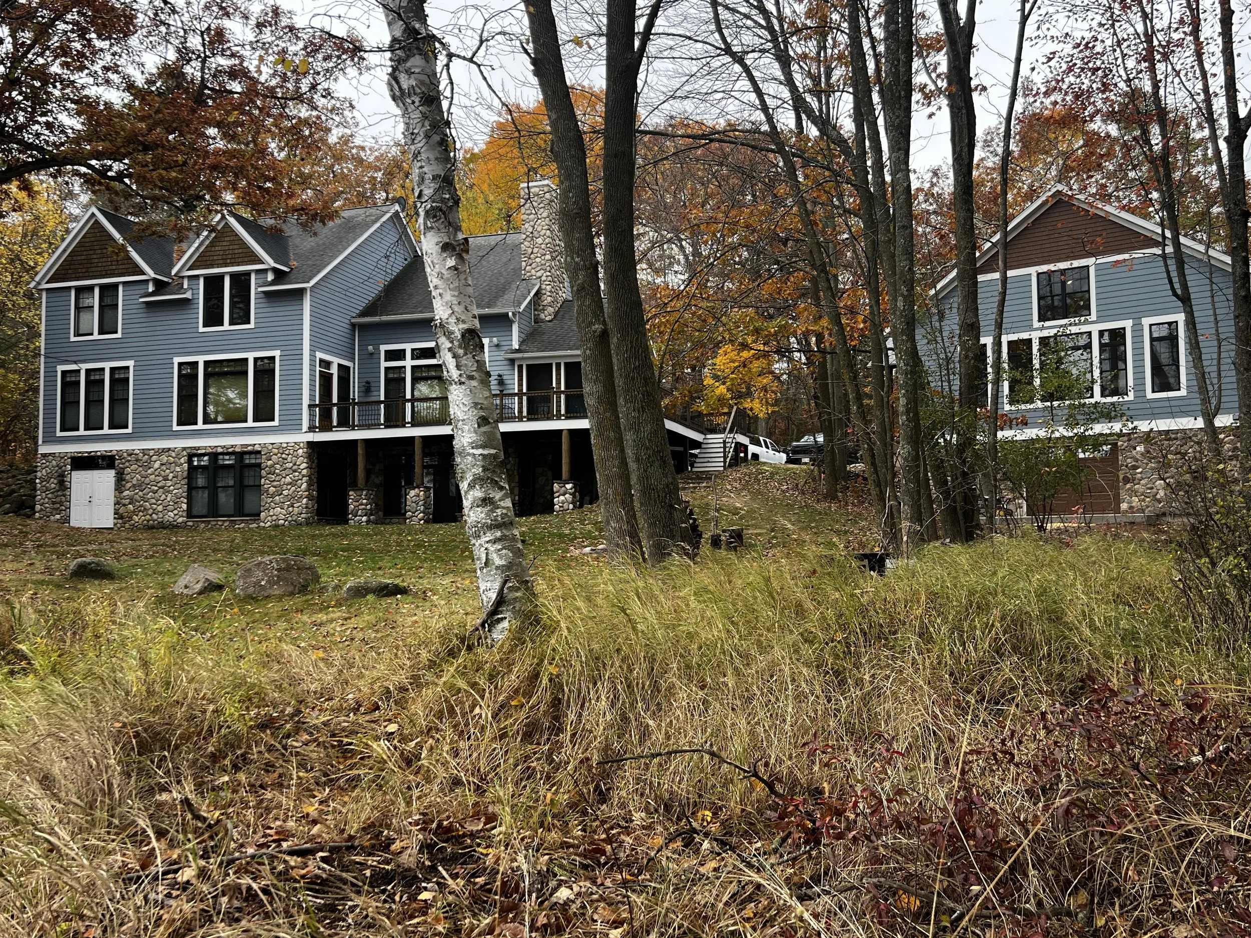 Lakeside home with blue siding and stone foundation surrounded by mature trees and autumn foliage.