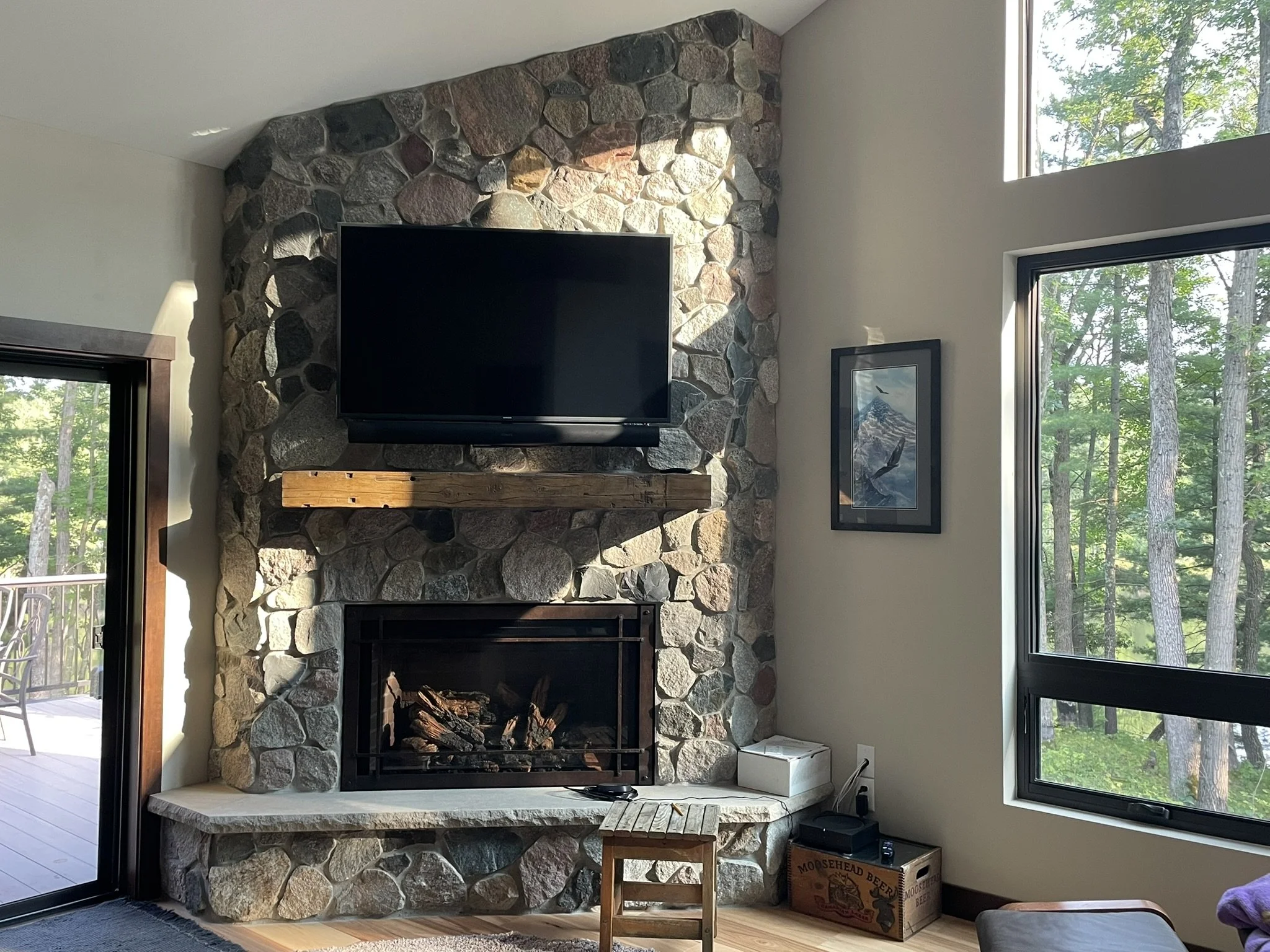 Living room stone fireplace with mounted TV, large windows, and natural light overlooking wooded surroundings.