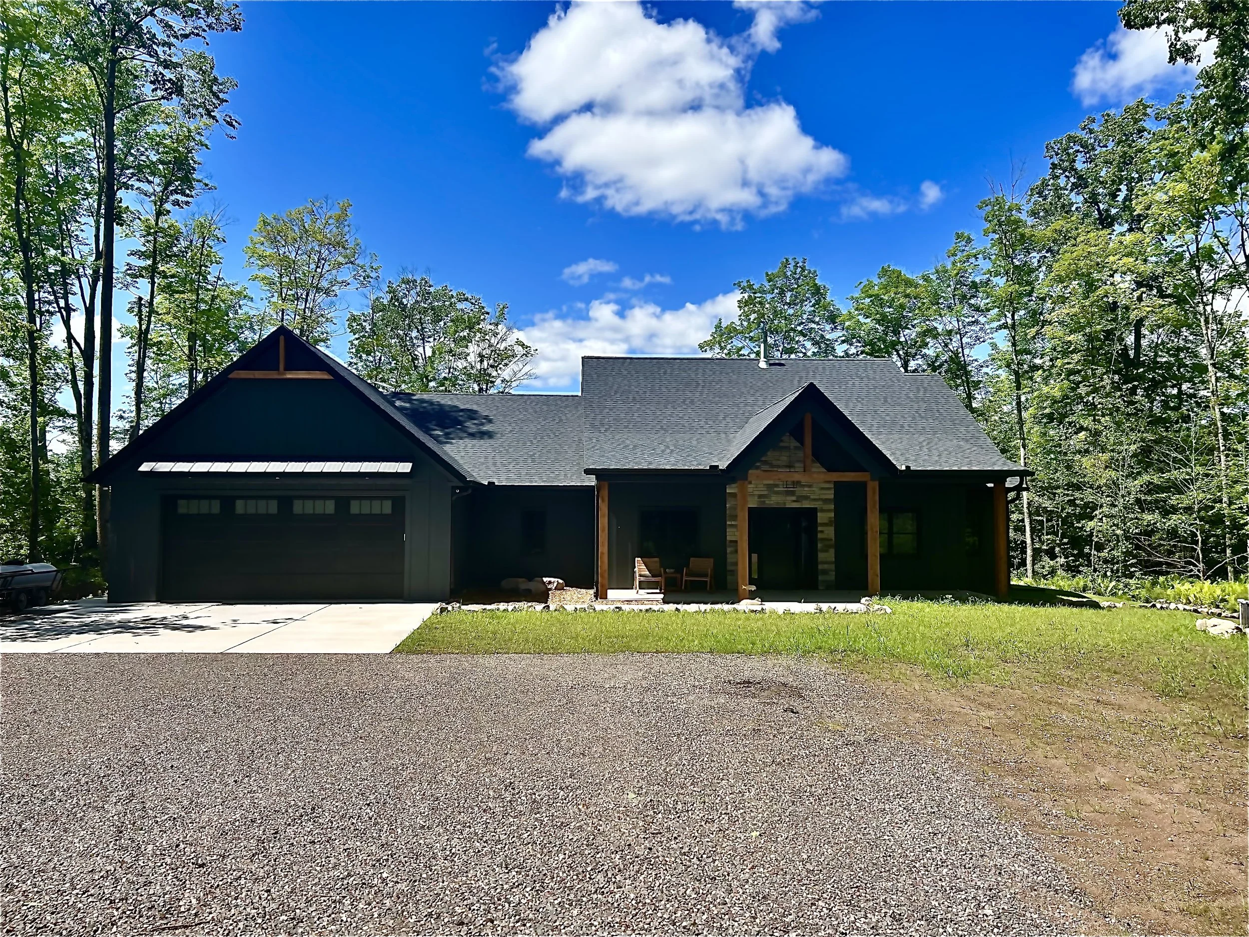 Modern craftsman-style home with dark siding, attached garage, covered porch, and wooded surroundings under a bright blue sky.